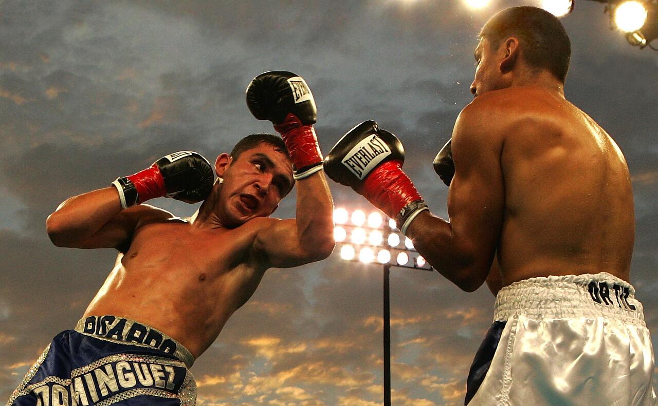 Dos boxeadores sin camiseta, con guantes y pantalones cortos, uno de ellos esquivando un puñetazo bajo las luces del estadio al atardecer.