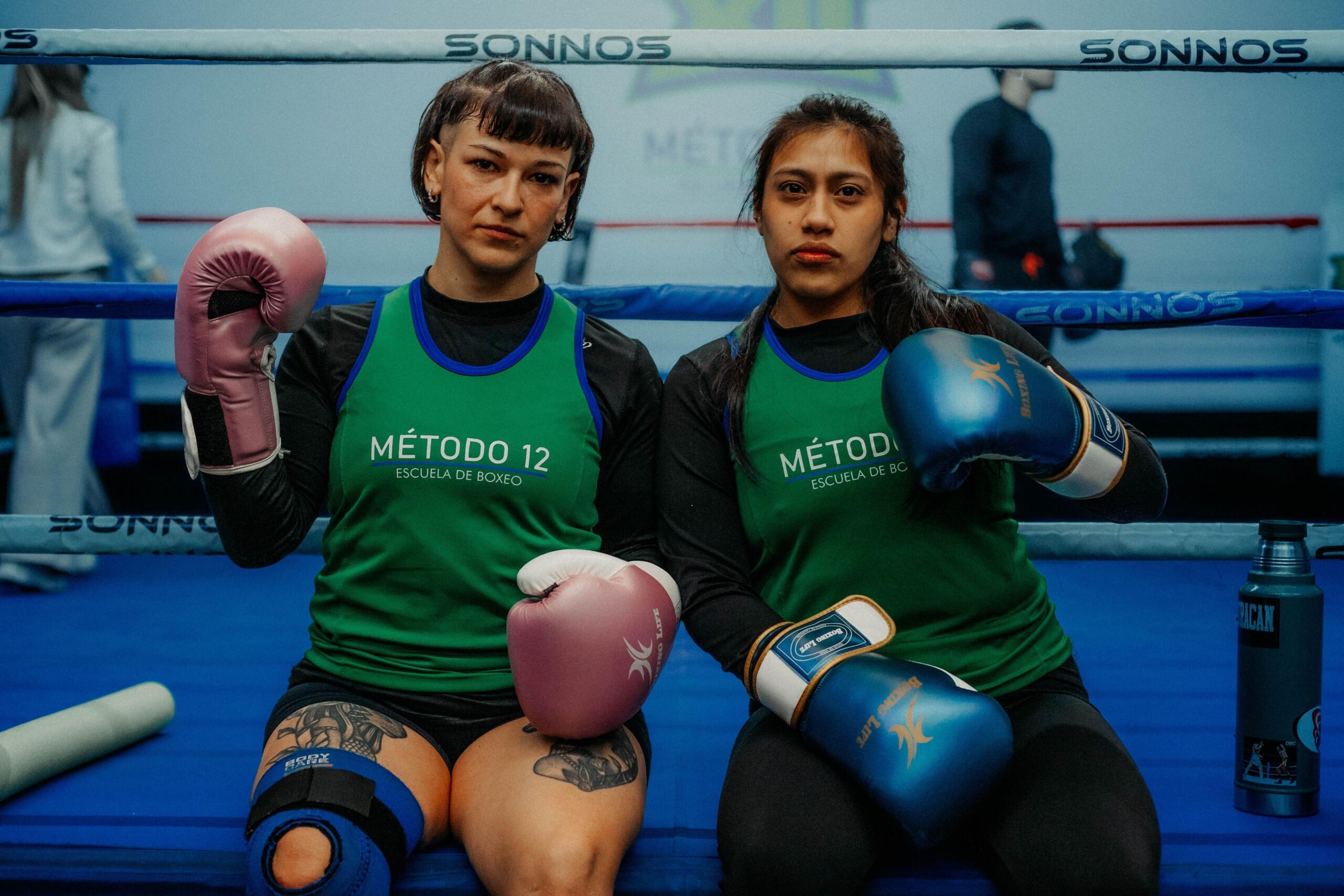 Dos mujeres con guantes de boxeo están sentadas en un ring de boxeo con camisetas verdes de «Método 12 Escuela de Boxeo»; una lleva guantes rosas y la otra, azules.