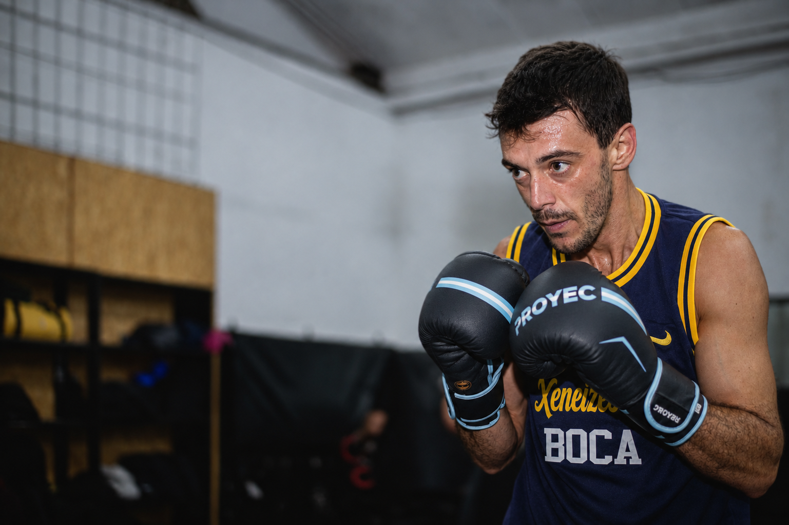 Un hombre con una camiseta de baloncesto azul marino con ribetes amarillos y guantes de boxeo negros, entrenando en un recinto cerrado con una postura concentrada.