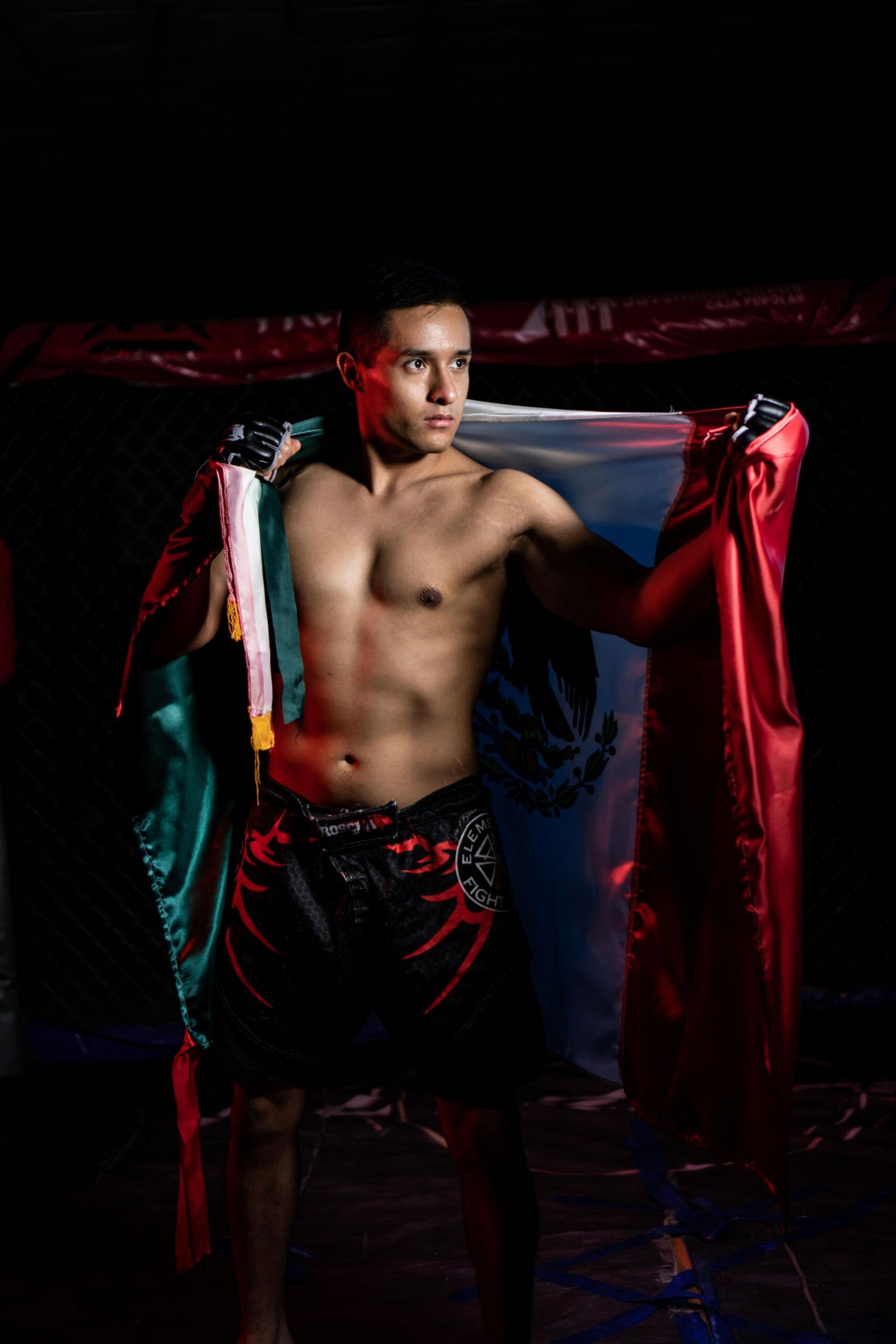Luchador sin camiseta, con pantalones cortos negros, que sostiene una bandera mexicana a la espalda, de pie en una jaula de combate con poca luz.