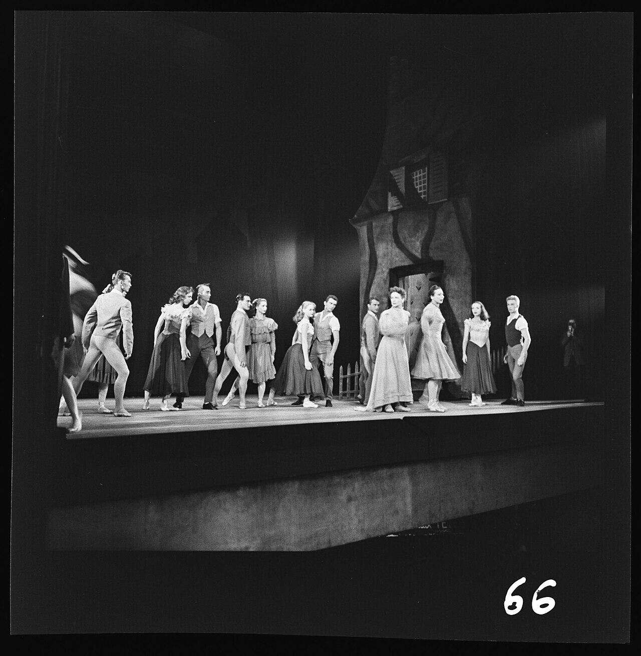 Fotografía en blanco y negro de un grupo de bailarines de ballet con sus trajes, actuando en el escenario frente a un decorado que representa una casa.