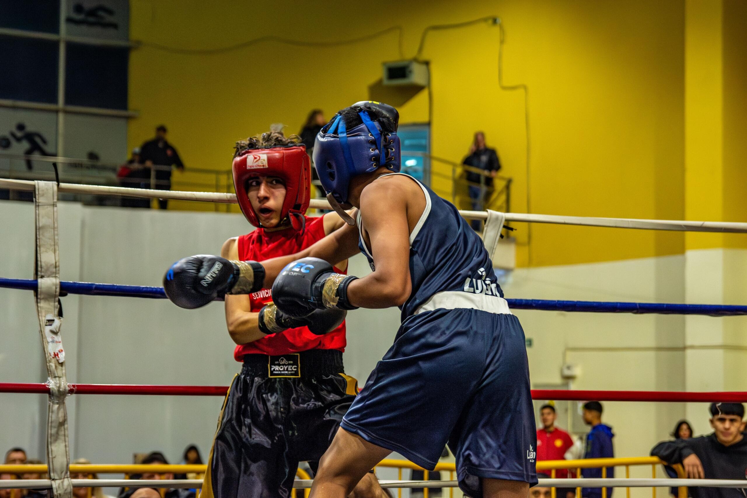 Dos jóvenes boxeadores, uno vestido de rojo y otro de azul, entrenando en un ring de boxeo con espectadores al fondo.