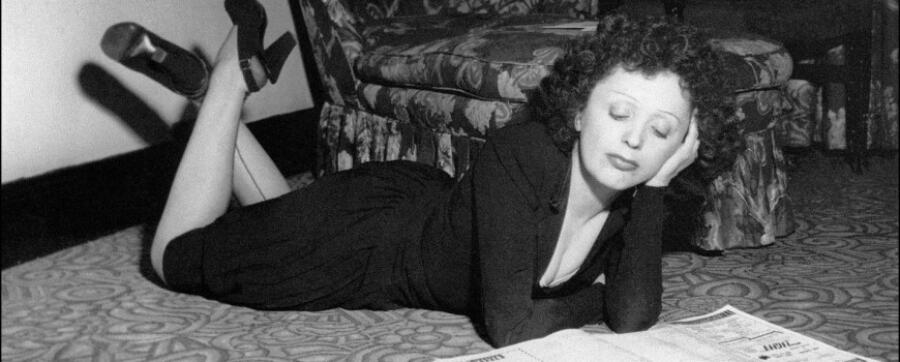 Una mujer de pelo rizado está recostada sobre la alfombra, apoyada en los codos, leyendo un periódico. Lleva un vestido oscuro, lo que transmite un estado de ánimo tranquilo e introspectivo.