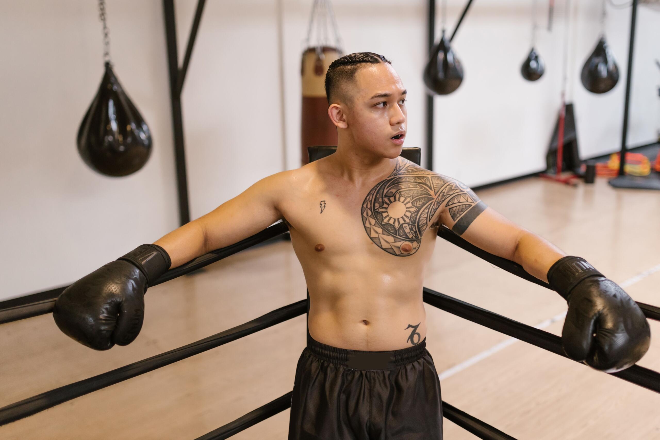Boxeador con tatuajes y guantes negros, apoyado en las cuerdas del ring, mirando hacia un lado en un gimnasio con sacos de boxeo al fondo.