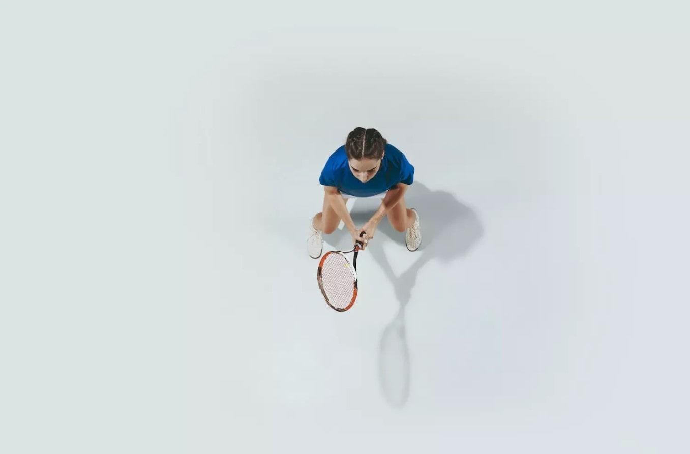 Vista desde arriba de una mujer con una camiseta azul que sostiene una raqueta de tenis, a punto de golpear una pelota, sobre un fondo blanco liso.