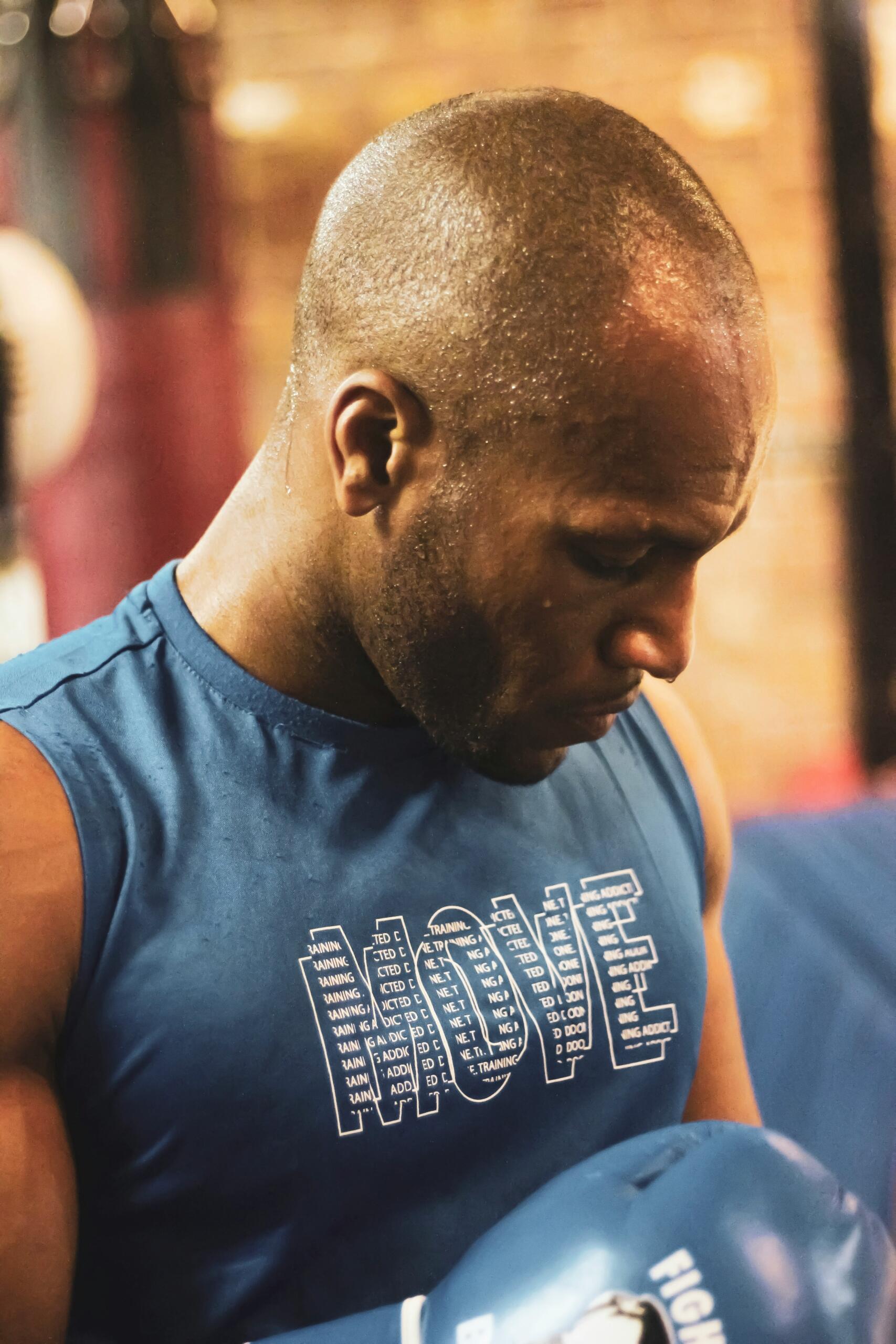 Una persona que lleva una camiseta azul sin mangas con la palabra «MOVE» impresa mira hacia abajo mientras sostiene un guante de boxeo azul en un gimnasio.