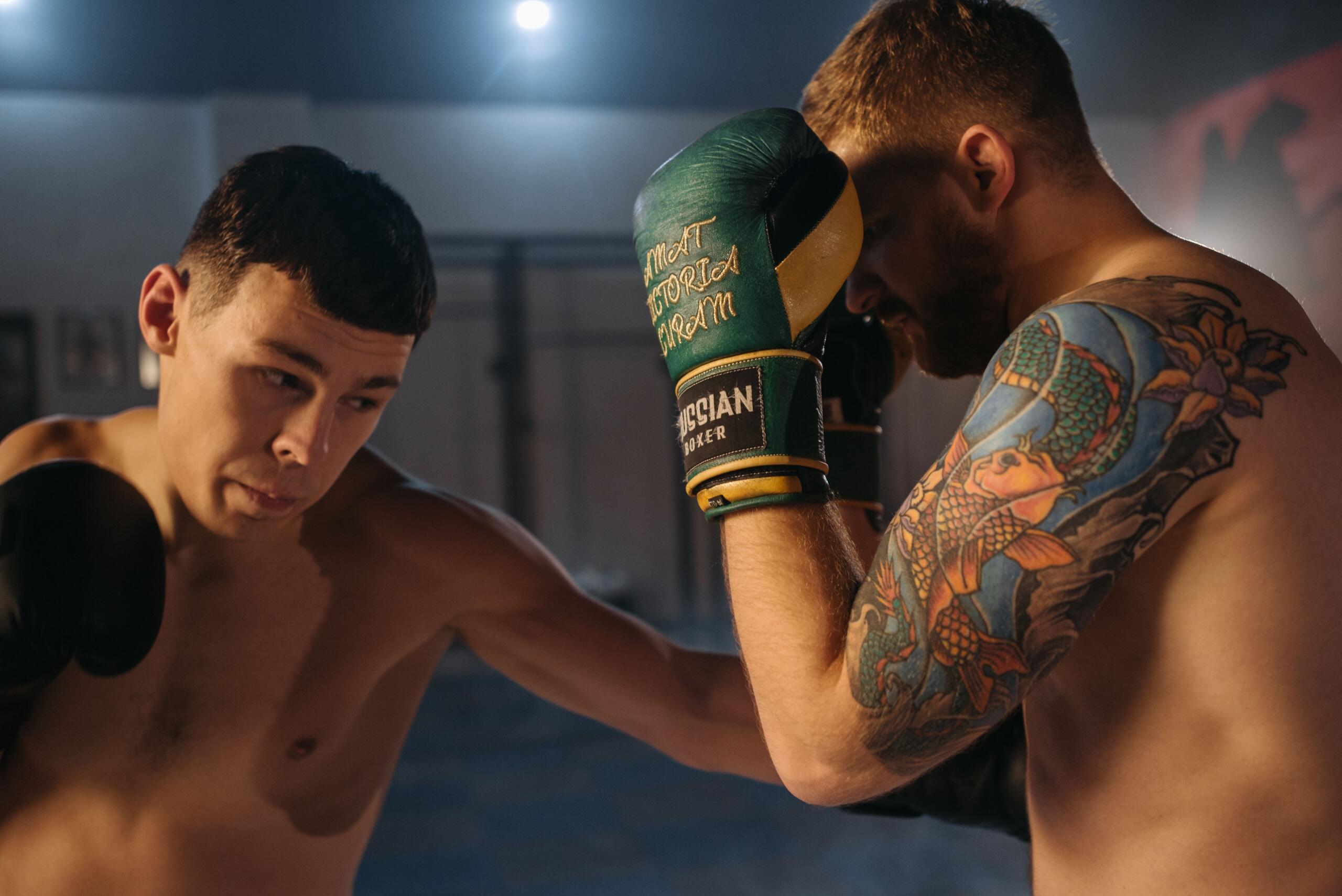 Dos boxeadores sin camiseta entrenan en un gimnasio con poca luz; uno lanza un puñetazo mientras el otro se defiende con los guantes y luce un brazo tatuado de colores vivos.