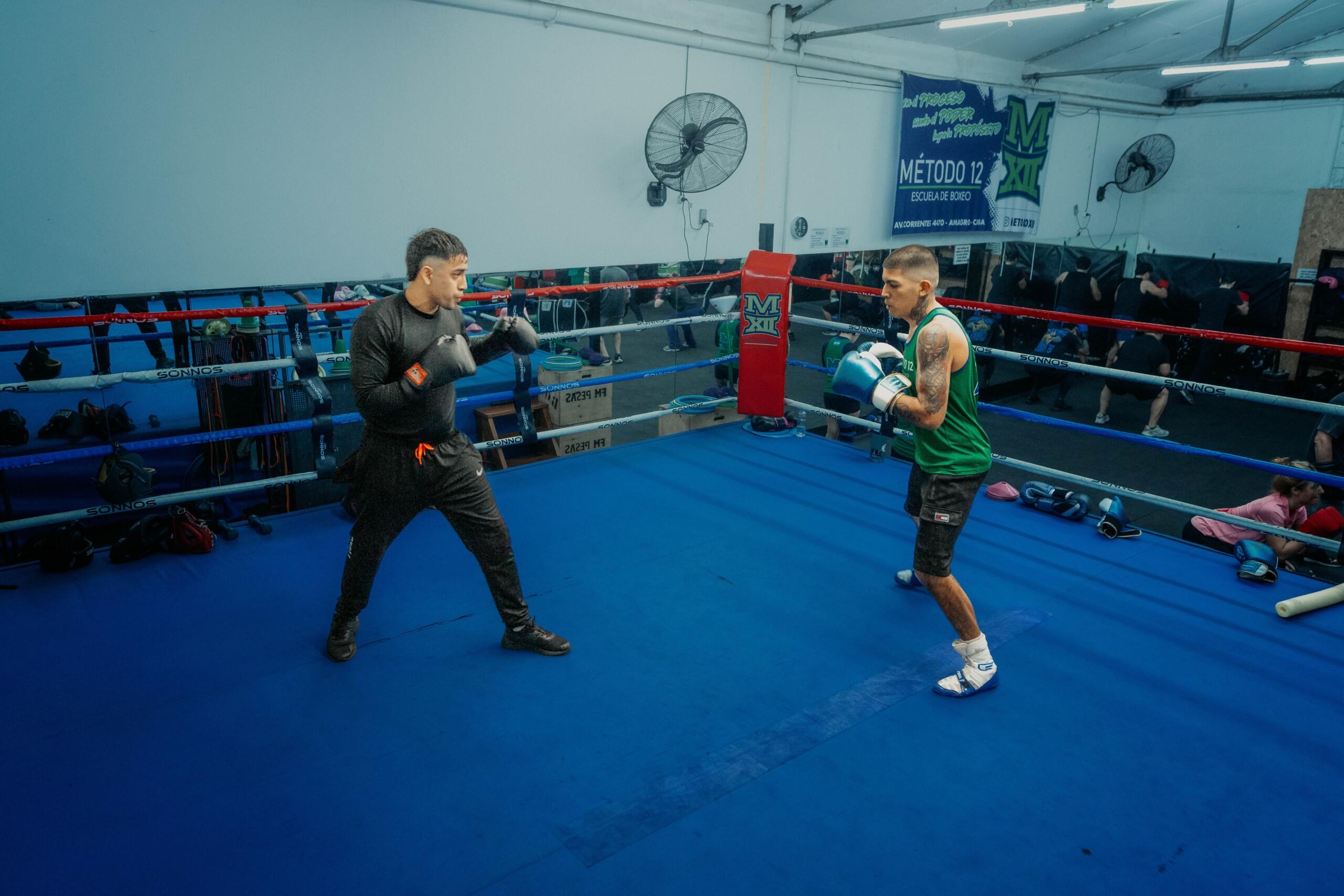 Dos boxeadores masculinos entrenan en un ring azul dentro de un gimnasio rodeado de espejos, aficionados y material de entrenamiento.