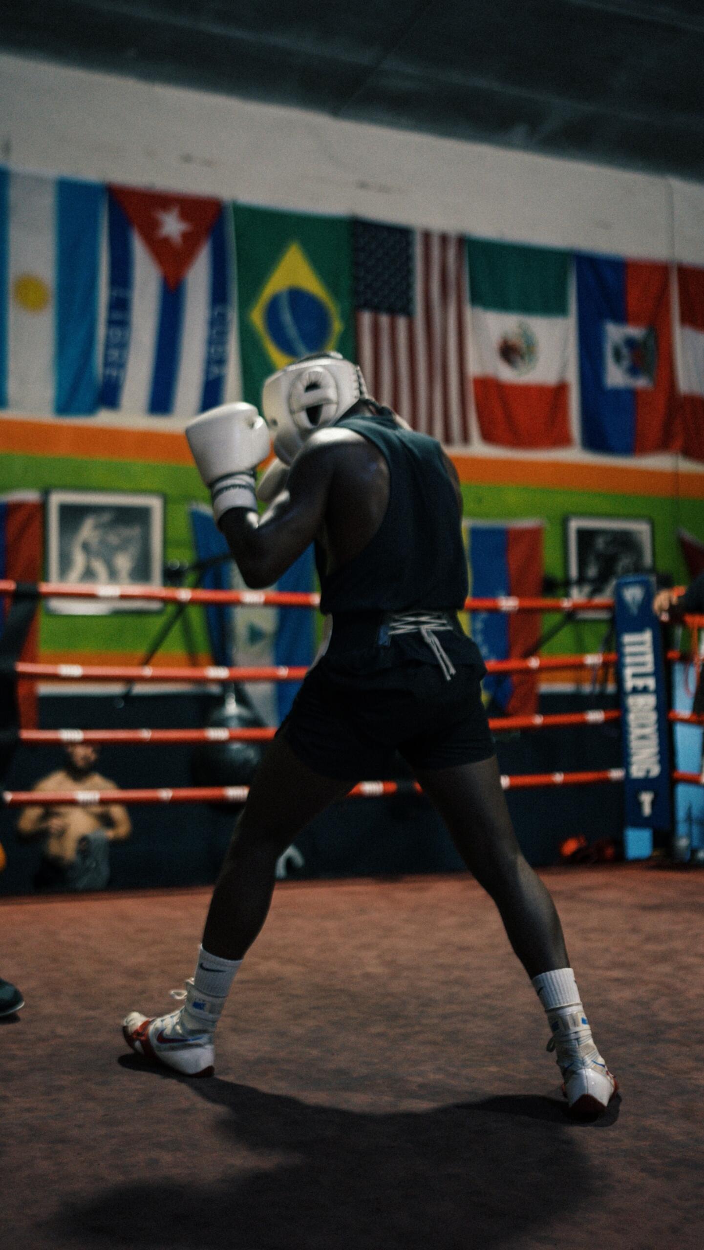 Boxeador con equipo de protección entrenando en un ring de gimnasio, con banderas internacionales y espectadores al fondo.