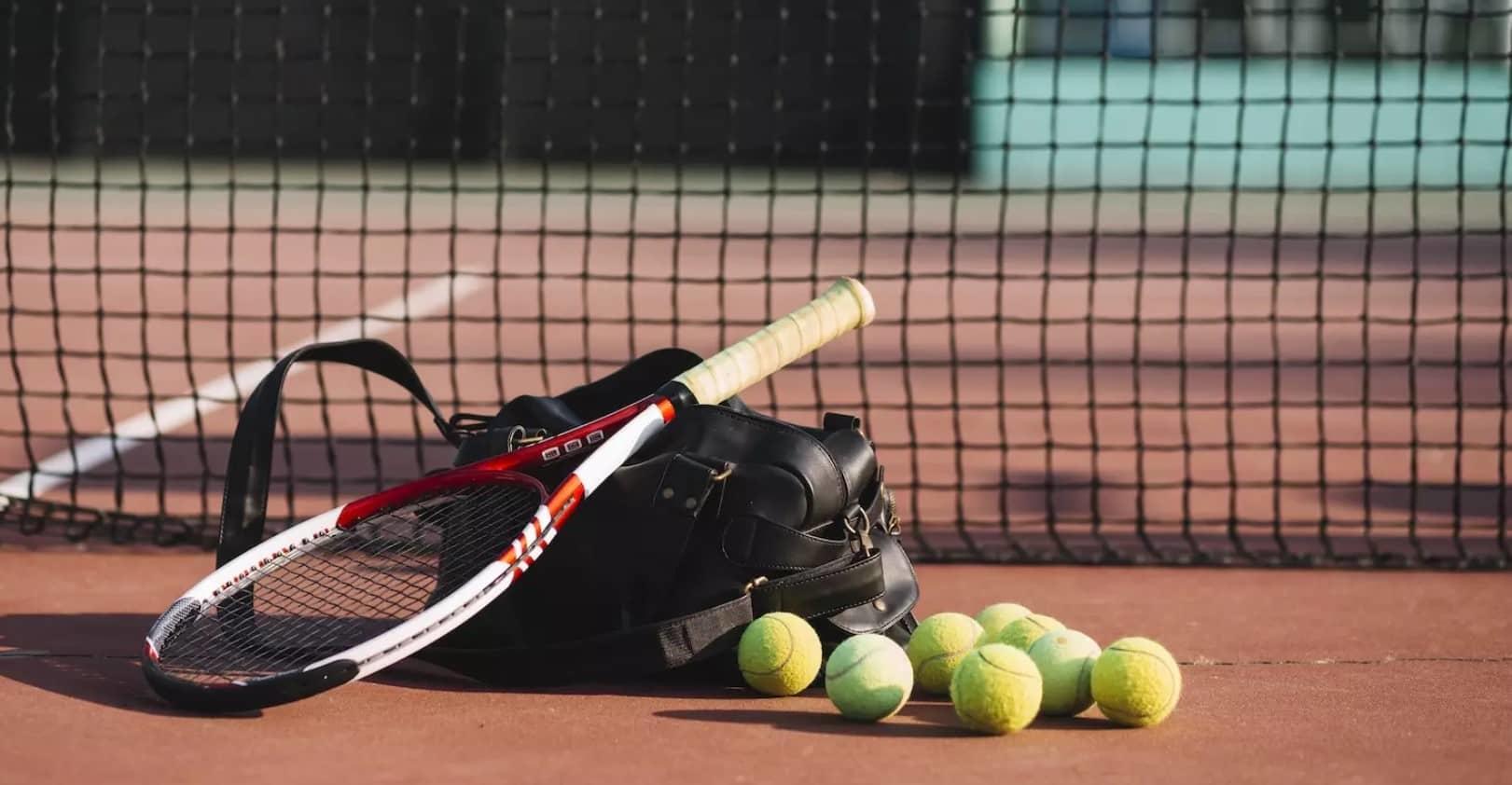 Raquetas de tenis apoyadas sobre una bolsa negra, con pelotas de tenis esparcidas por una pista de tierra batida cerca de la red.