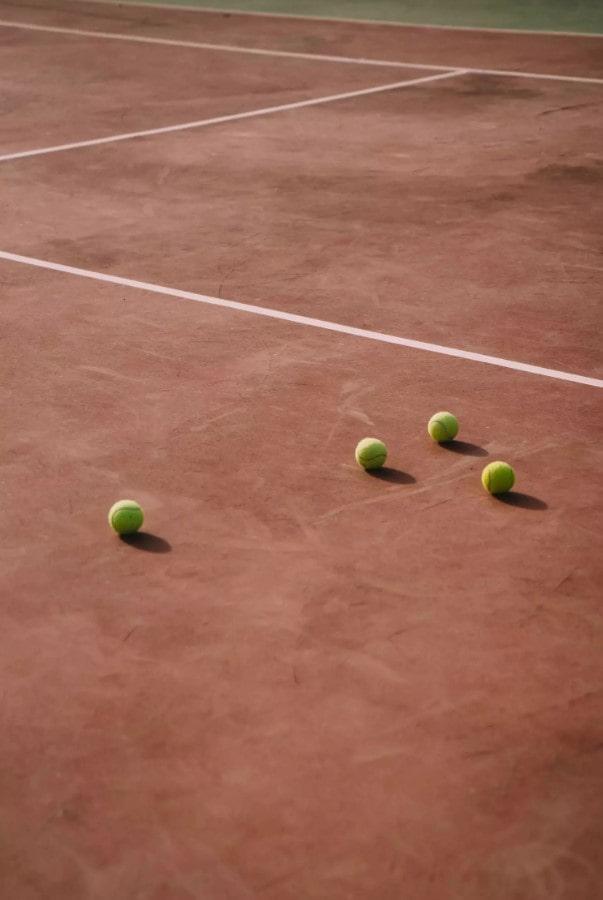 Cuatro pelotas de tenis esparcidas por una pista de tierra batida con líneas de delimitación blancas.