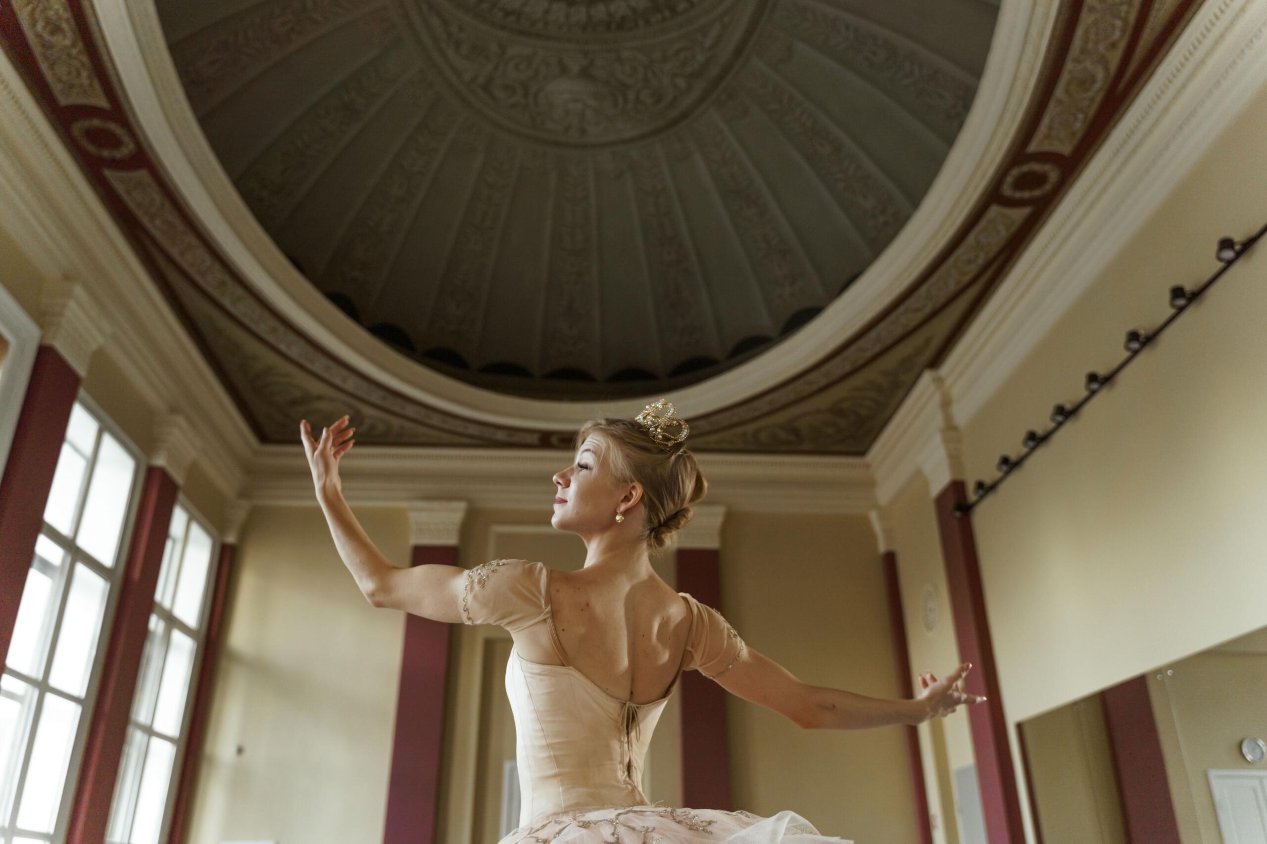 Una bailarina con una tiara y un vestido de ballet que baila con elegancia en una amplia sala con ventanas altas y un techo abovedado ornamentado.