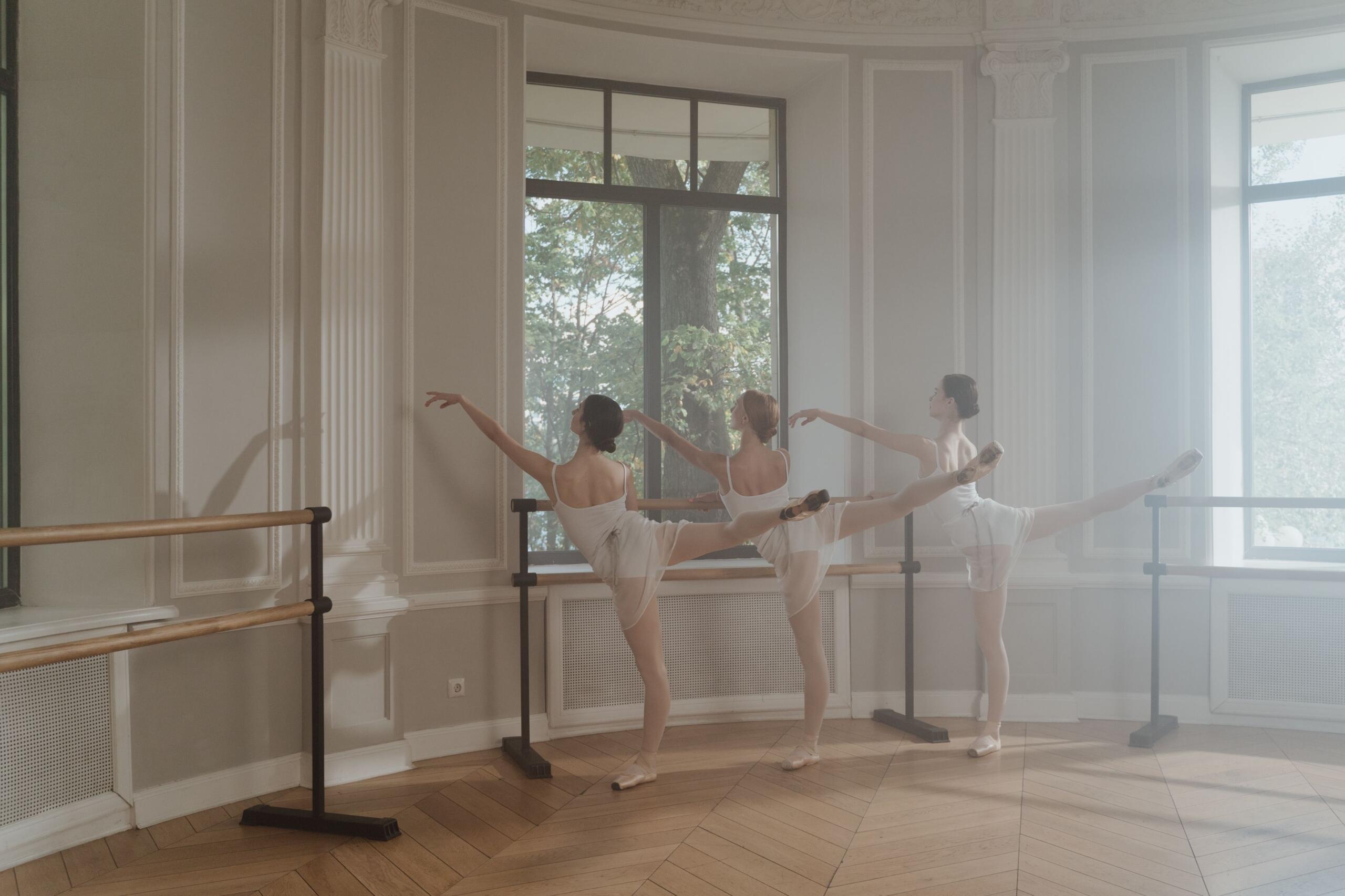 Tres bailarinas vestidas de blanco practican una pose de arabesco junto a una barra en un elegante estudio de danza bañado por la luz del sol y con grandes ventanales.