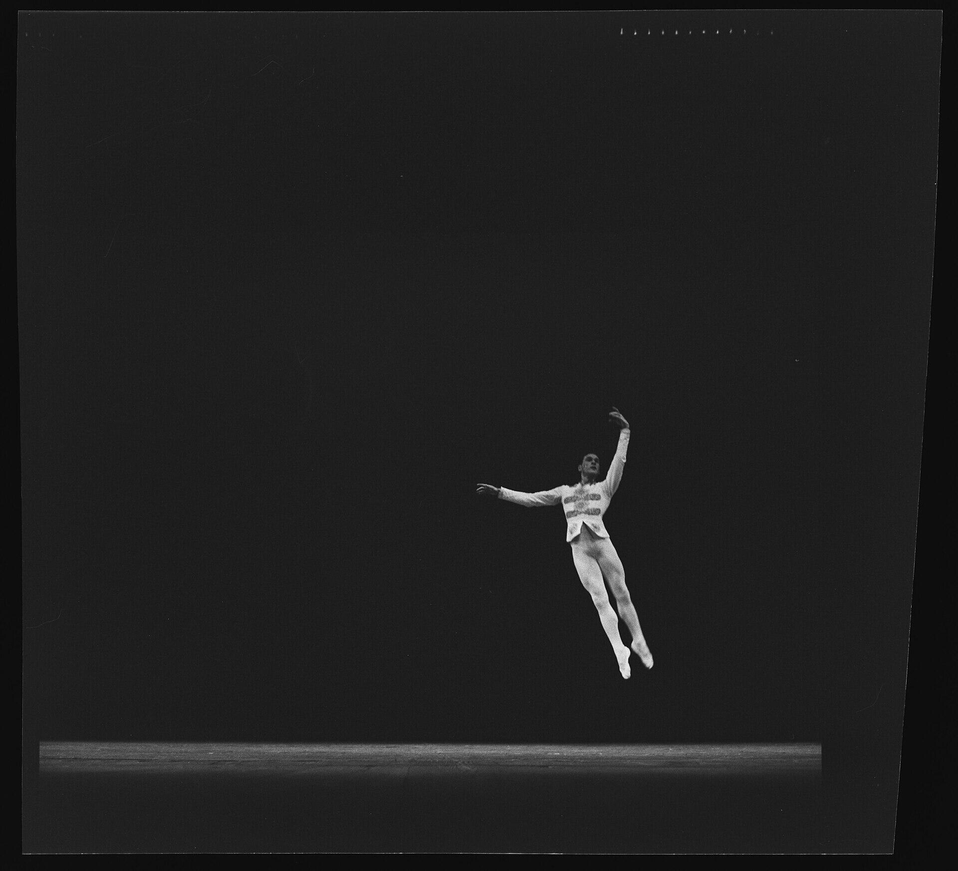 Un bailarín de ballet masculino capturado en el aire durante un salto potente y elegante, con el cuerpo inclinado y las piernas extendidas, un brazo hacia el lado y el otro arqueado sobre la cabeza, vistiendo una chaqueta de traje de ballet ornamentada de color blanco con mallas a juego, sobre un escenario oscuro con un fondo negro vacío, en una fotografía histórica en blanco y negro.