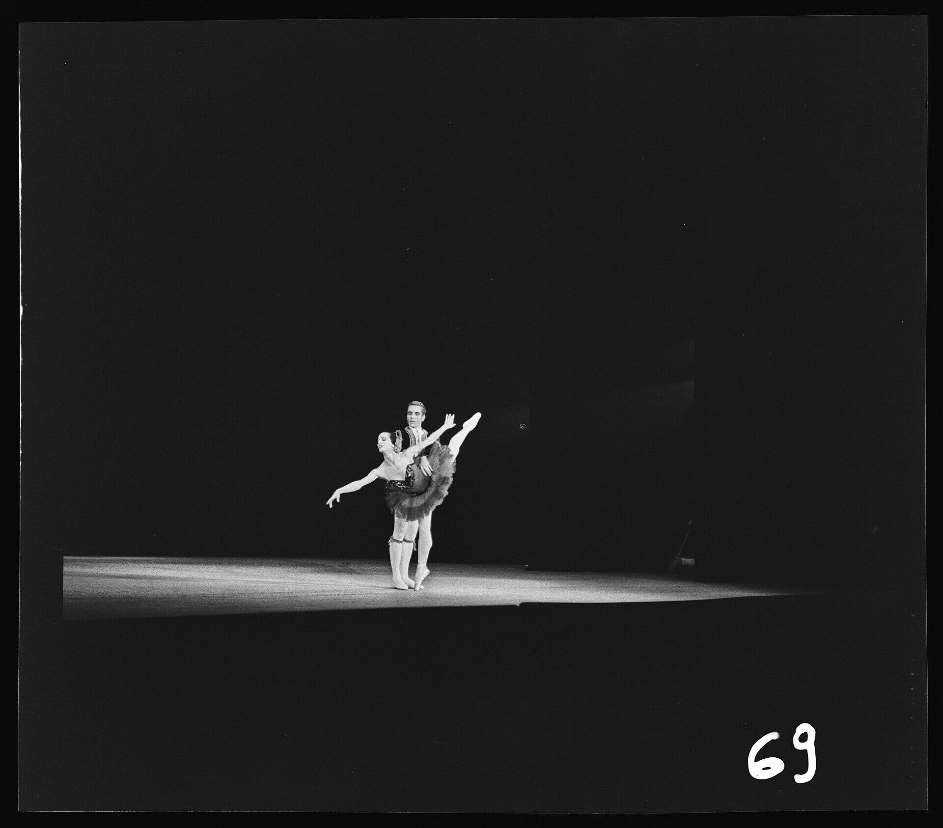 Fotografía histórica en blanco y negro de una pareja de bailarines ejecutando un pas de deux sobre un escenario iluminado. La bailarina sostiene una pose en puntas con la pierna elevada mientras es apoyada desde atrás por su compañero, en una escena representativa del origen y la historia del ballet clásico.