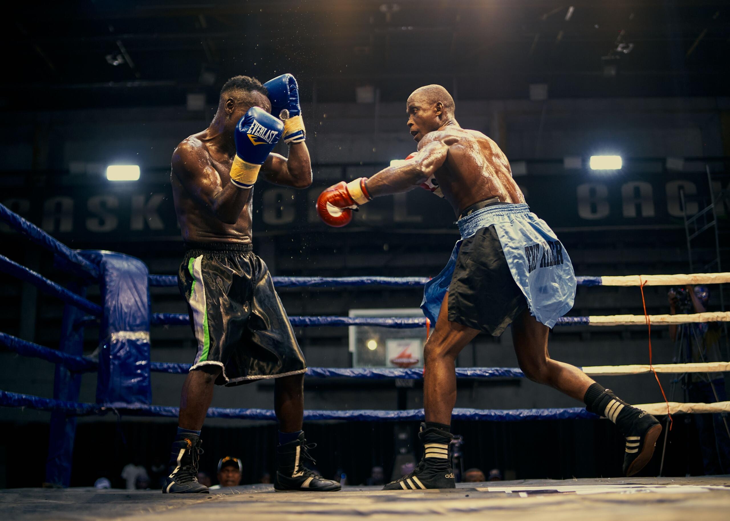 Dos boxeadores en un ring: uno se defiende con guantes azules y el otro lanza un puñetazo con guantes rojos bajo las brillantes luces del estadio.