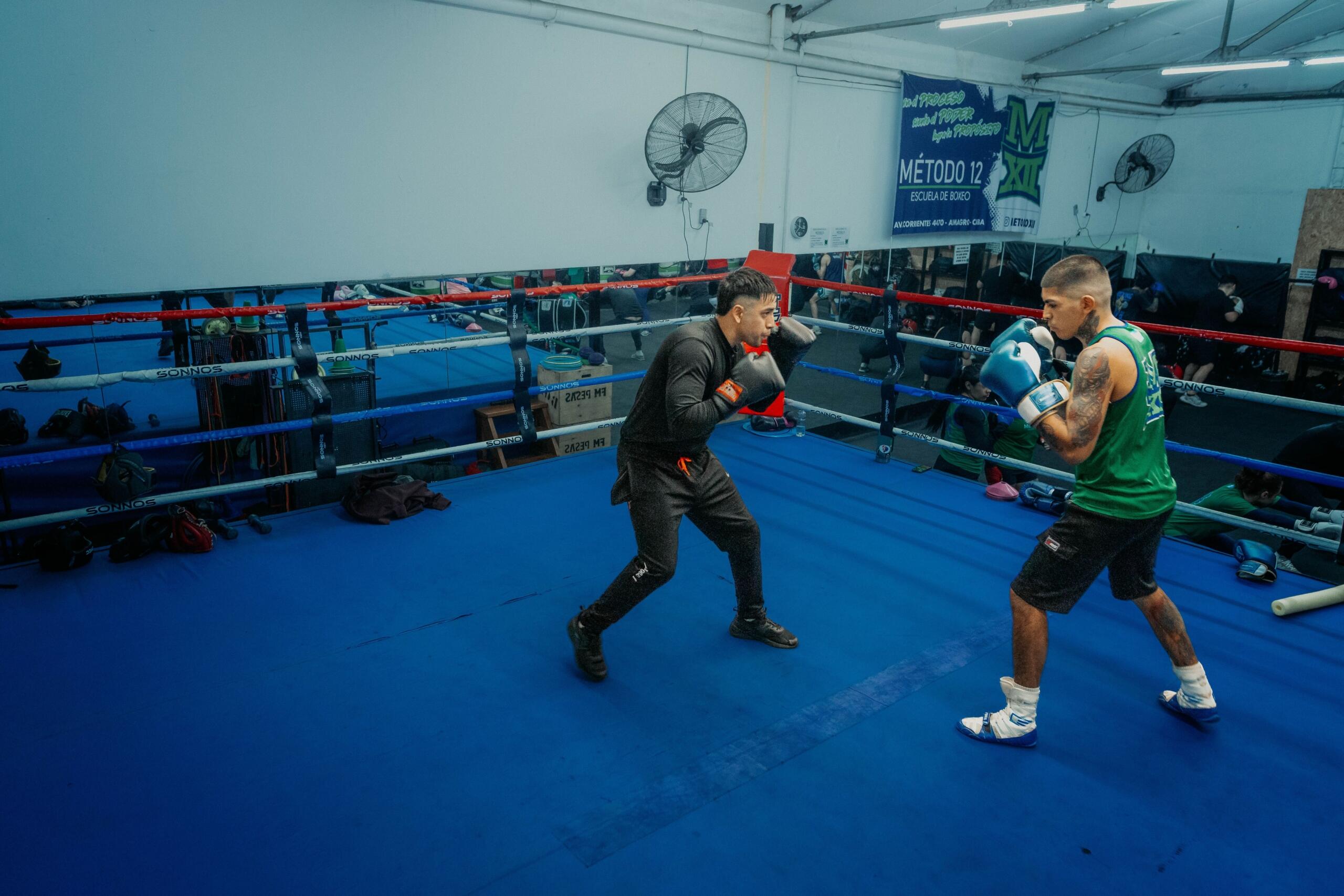 Dos boxeadores entrenando en un ring de boxeo con suelo azul y cuerdas rojas, blancas y azules, en un gimnasio con ventiladores de pared y un cartel.