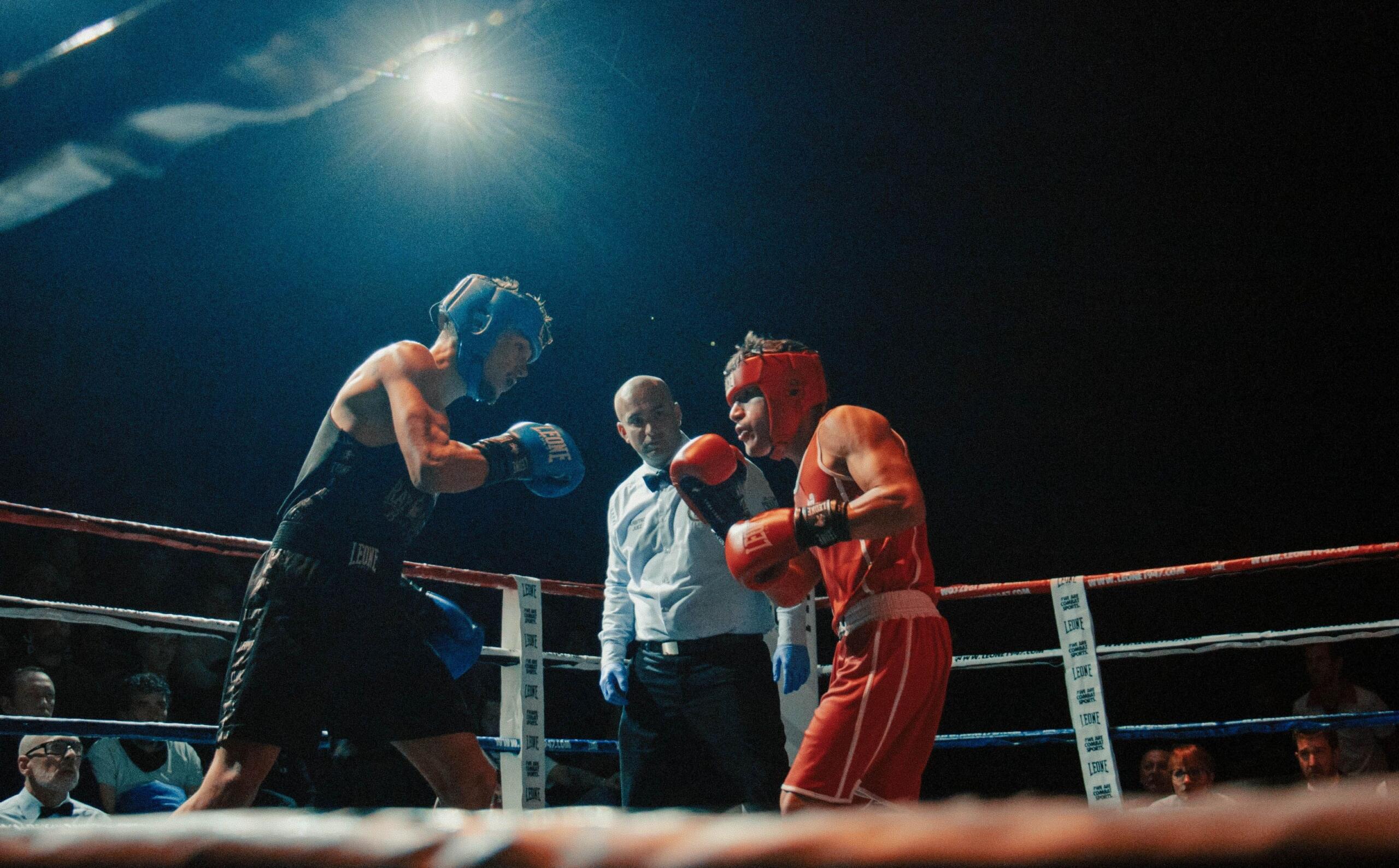 Dos boxeadores, equipados con cascos y guantes, se enfrentan en un ring bajo una intensa luz cenital, mientras un árbitro los observa atentamente.