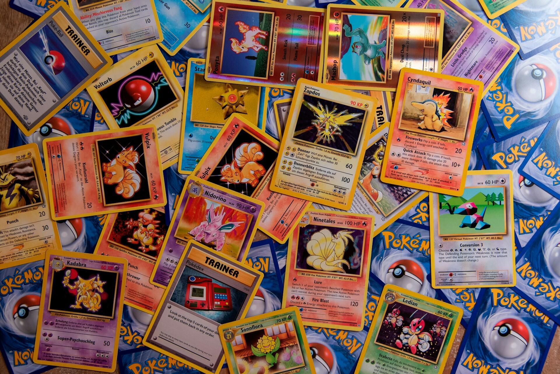 Una colección dispersa de diversas cartas coleccionables de Pokémon, entre las que se incluyen Vulpix, Zapdos, Cyndaquil y cartas de Entrenador, sobre una superficie de madera.