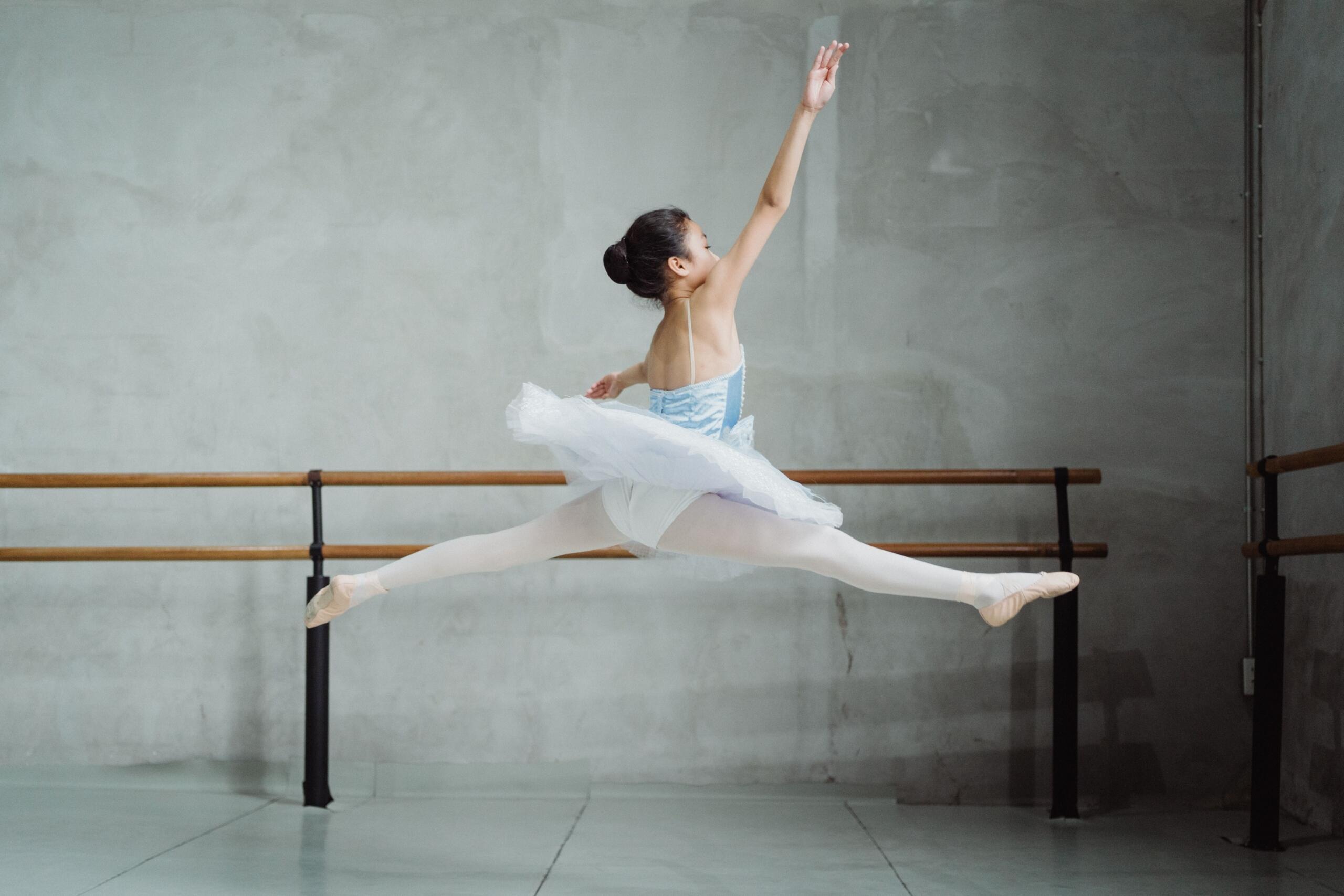 Una bailarina con un tutú blanco realizando un salto con apertura en el aire en un estudio de danza con barras de ballet y una pared gris de fondo.