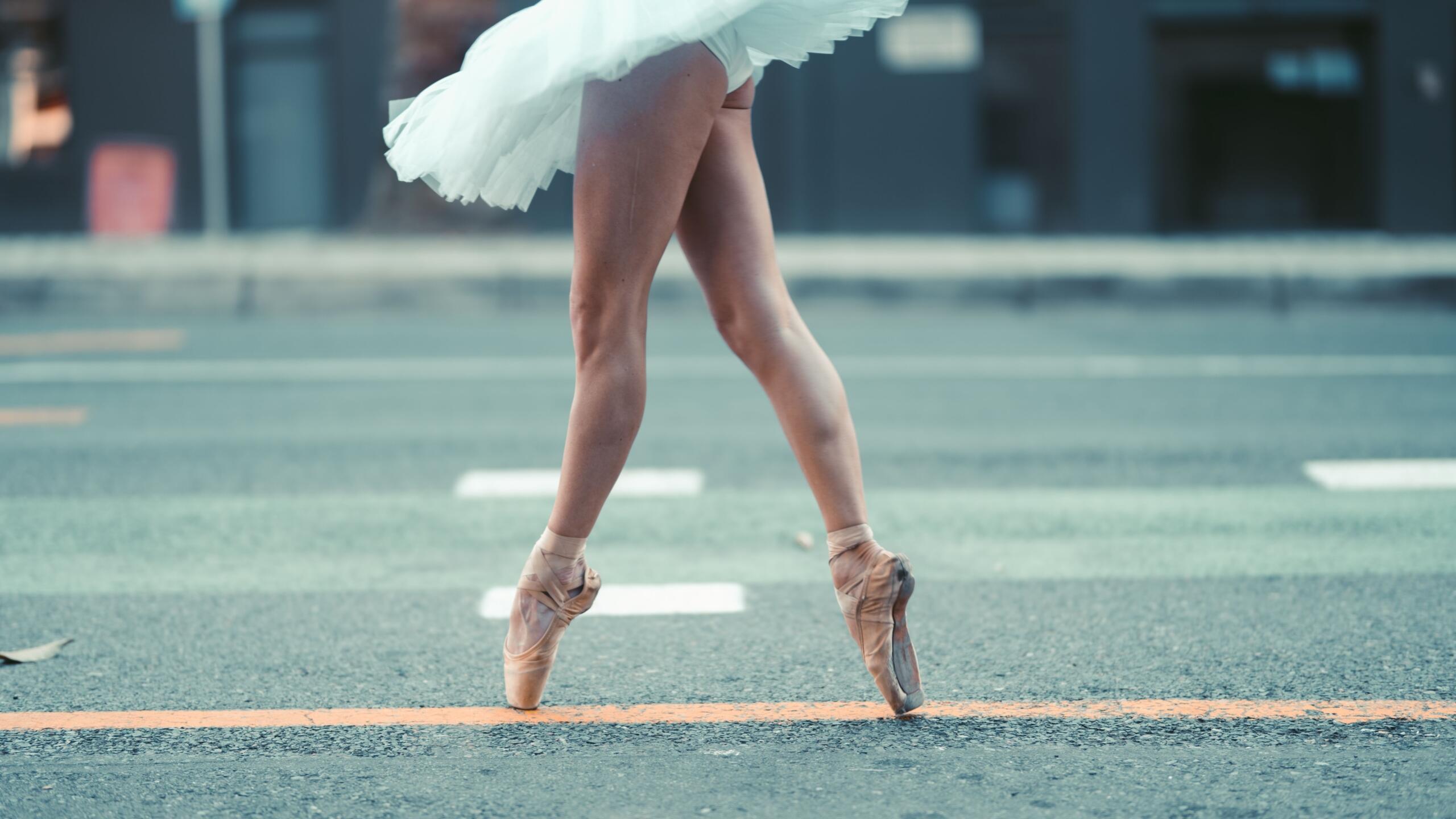 Una bailarina de ballet con zapatillas de punta y un tutú blanco, manteniendo el equilibrio en puntas en una calle con una línea naranja.