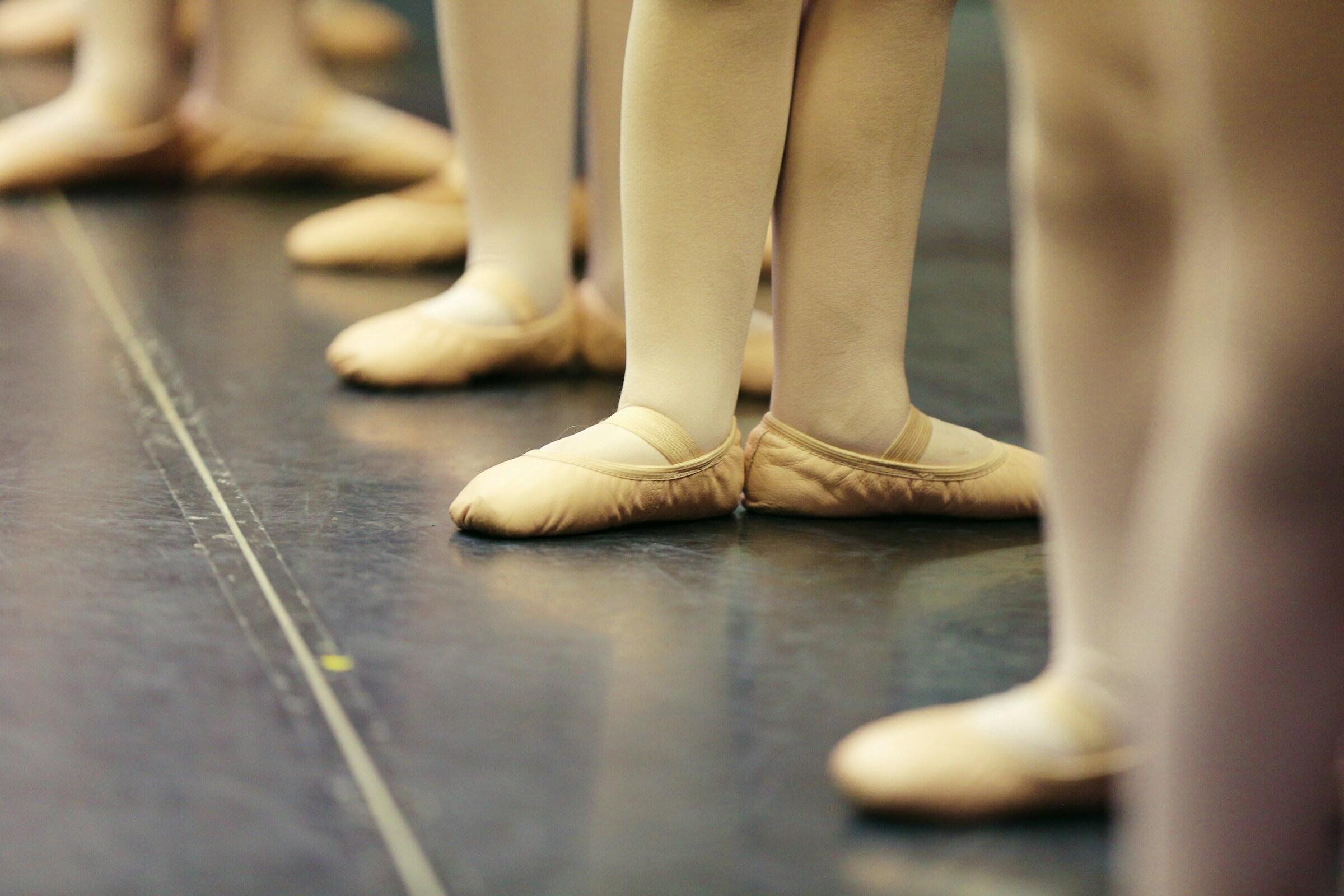 Primer plano de los pies de unas bailarinas de ballet con medias beige y zapatillas de ballet, alineados sobre el suelo negro del escenario.