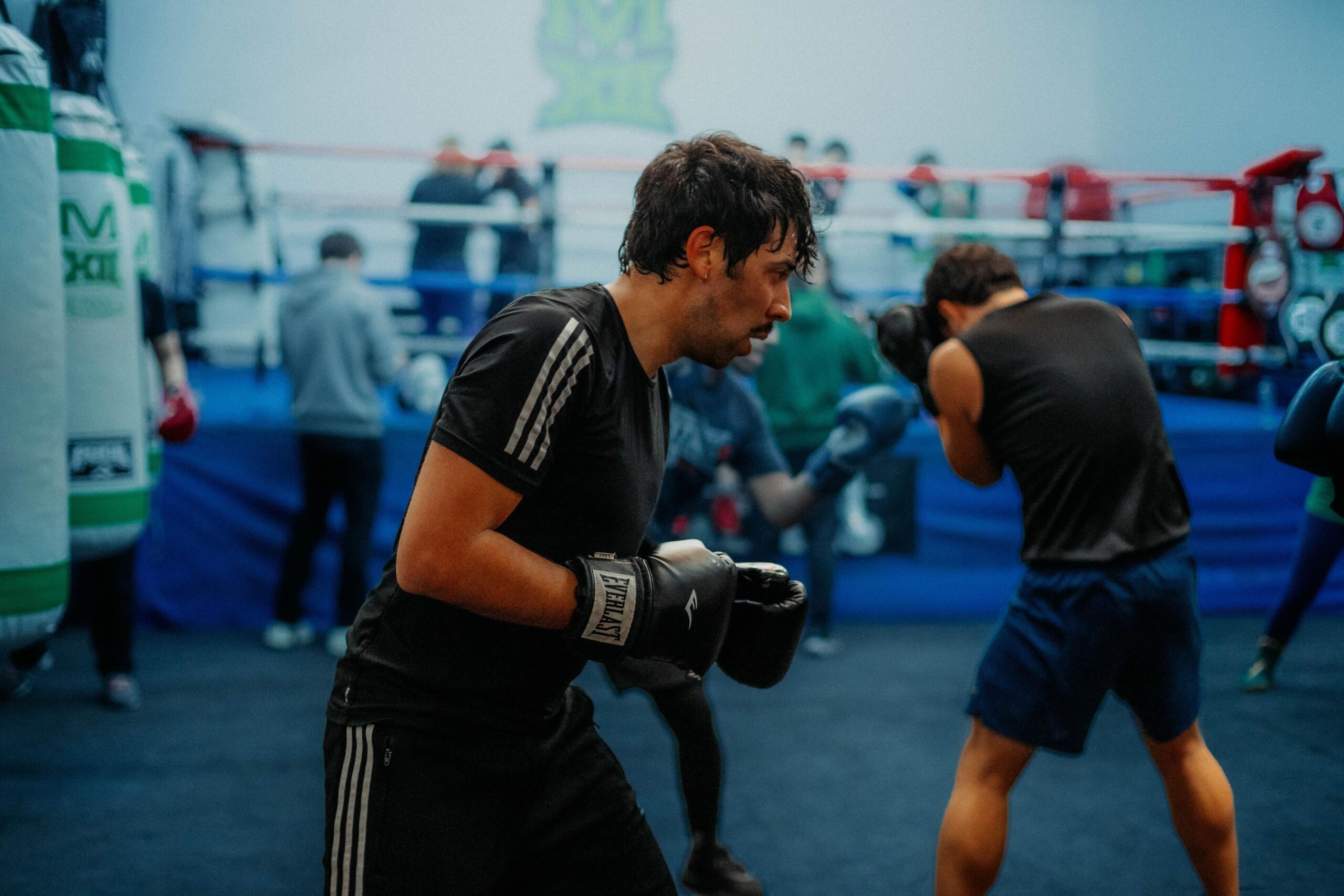 Dos hombres boxeando en un ring de gimnasio, con guantes y ropa deportiva, con otras personas y material de boxeo al fondo.