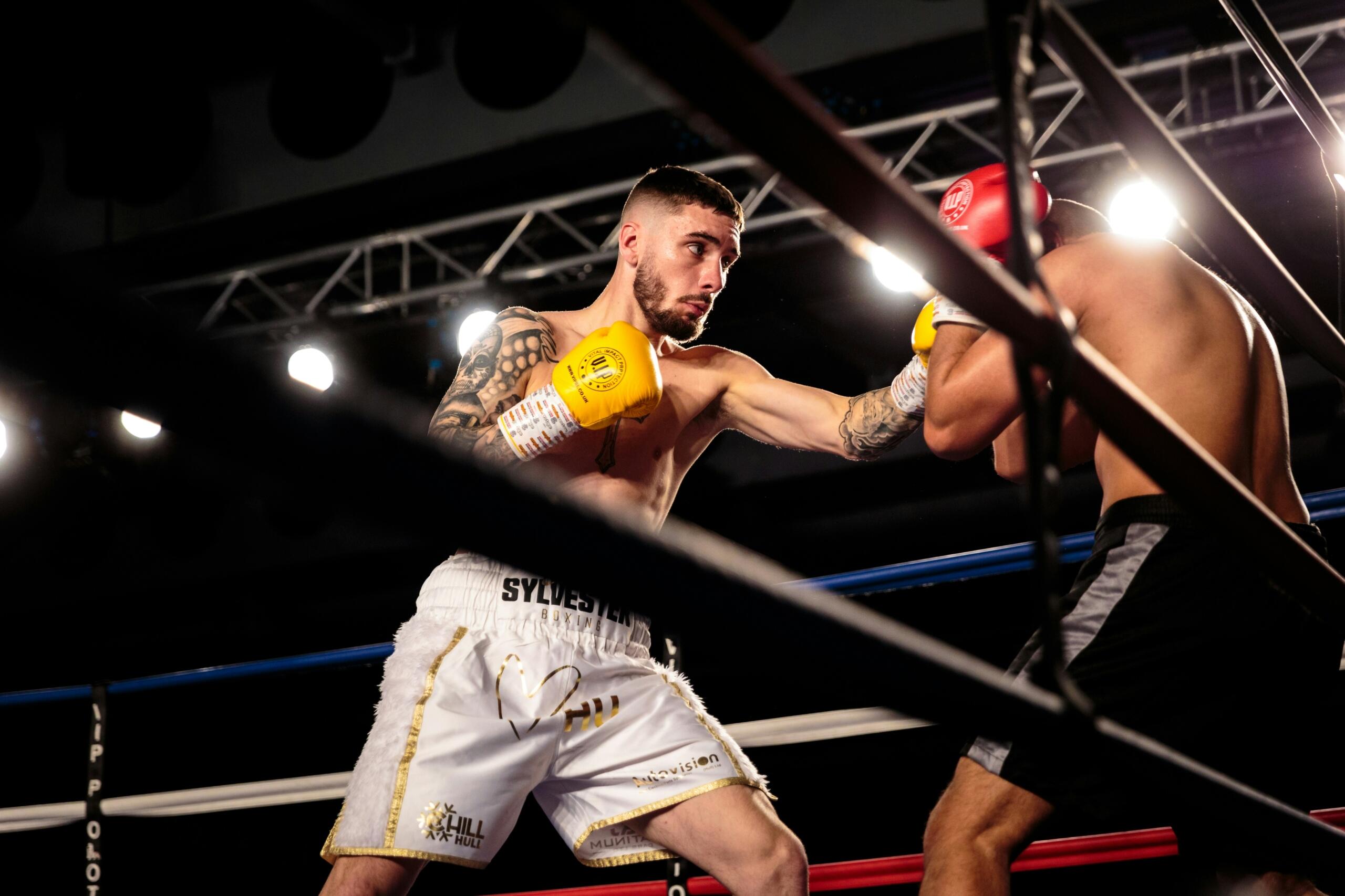Dos boxeadores en un ring, uno con guantes amarillos que ataca y el otro, con guantes rojos, que se defiende, bajo unas potentes luces de techo.