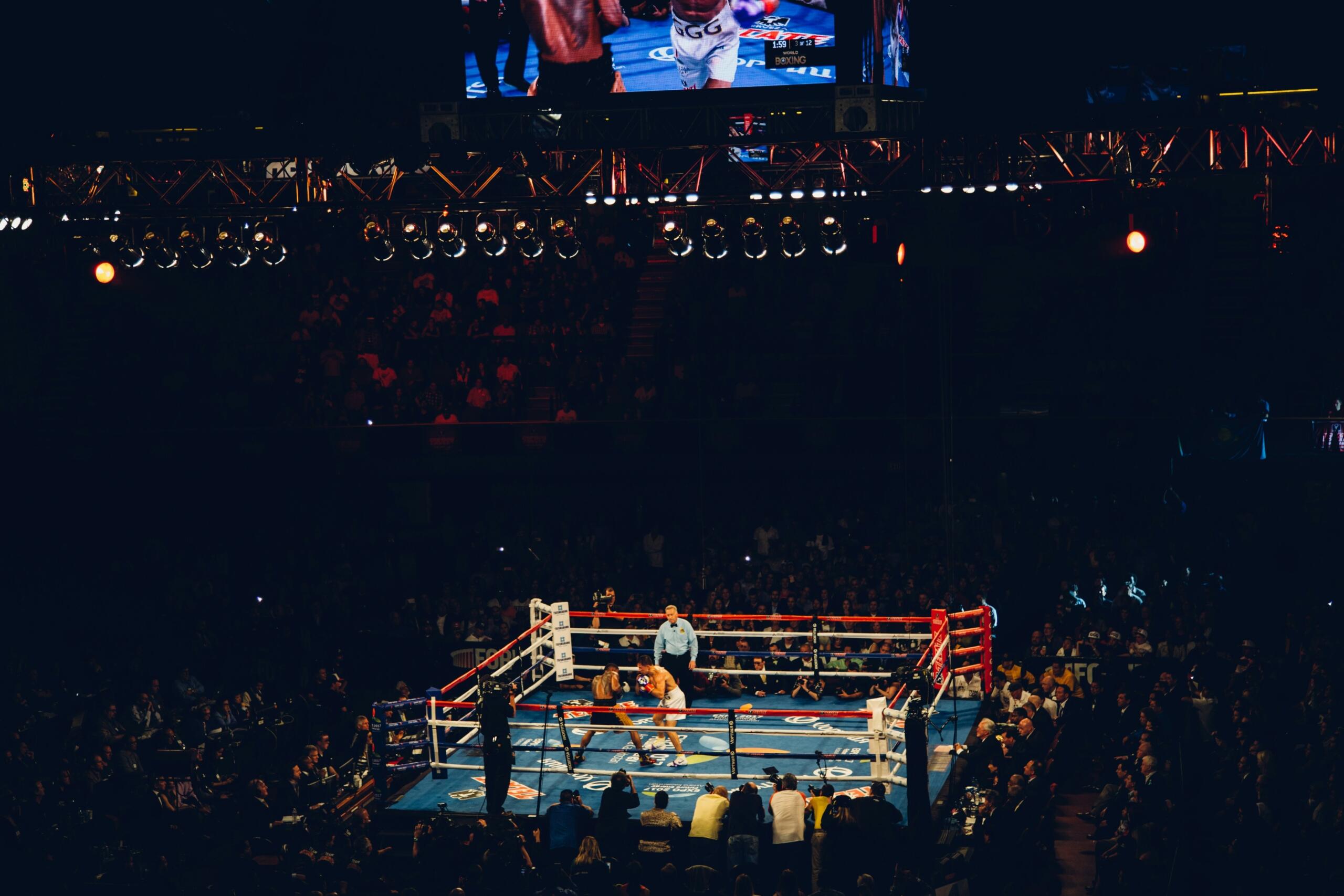 Combate de boxeo en un gran estadio con poca luz, con espectadores y un árbitro dentro del ring.