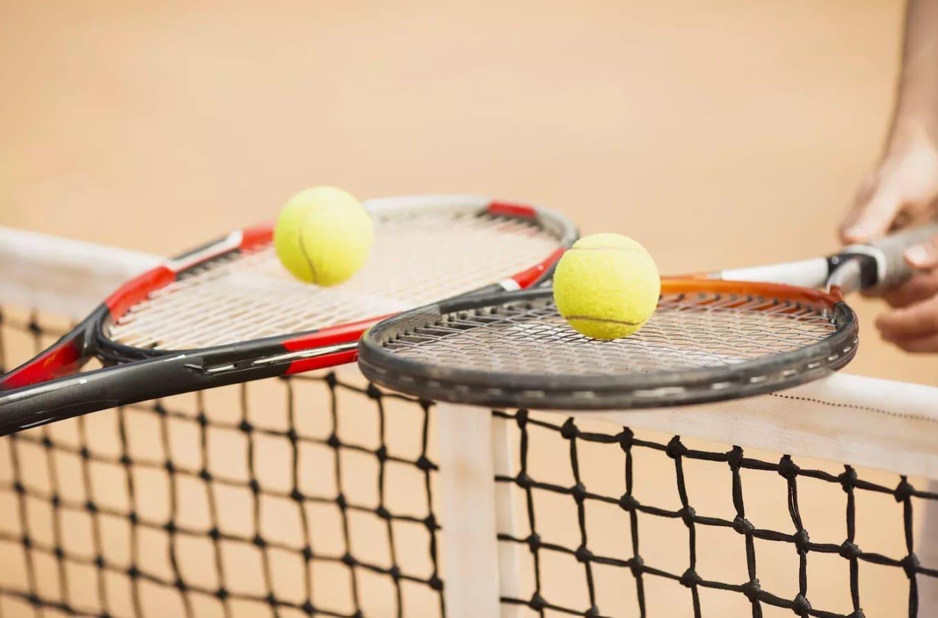 Dos raquetas de tenis con pelotas que rebotan en sus cuerdas por encima de una red de tenis en una pista de tierra batida, en la que se ve la mano de un jugador.