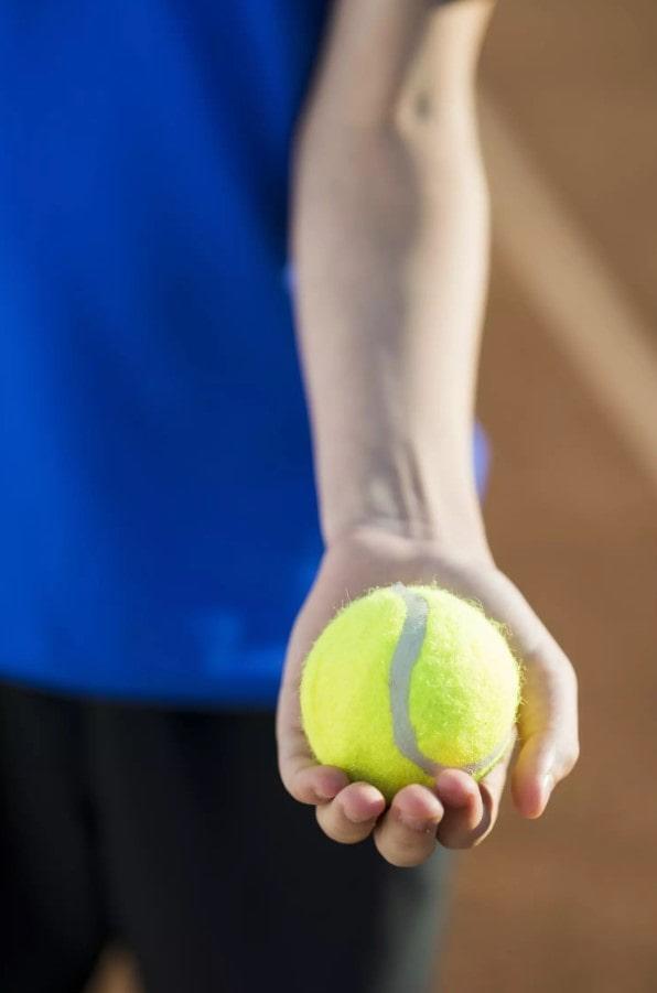 Primer plano de una persona que sostiene una pelota de tenis de color amarillo brillante en la mano, con una camiseta azul y un fondo desenfocado.