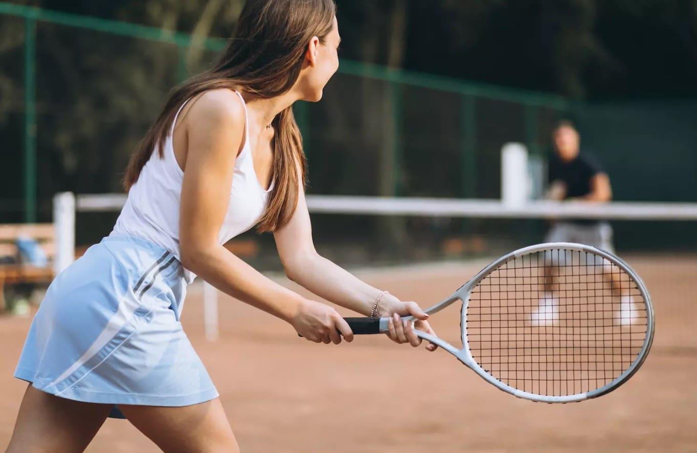 Mujer vestida con ropa deportiva, con una raqueta de tenis en la mano, en una pista de tierra batida, preparándose para devolver un golpe durante un partido.