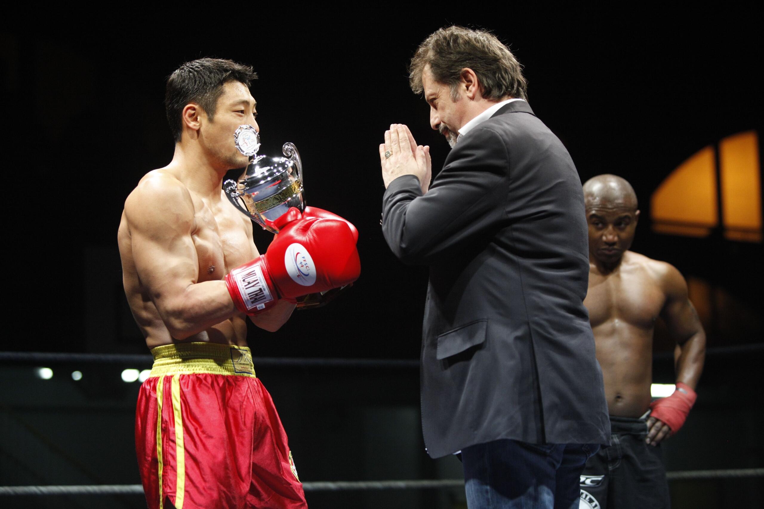 Un boxeador con guantes y pantalones cortos rojos sostiene un trofeo, mientras un hombre trajeado le hace un gesto de respeto y otro boxeador observa la escena en un ring con poca luz.