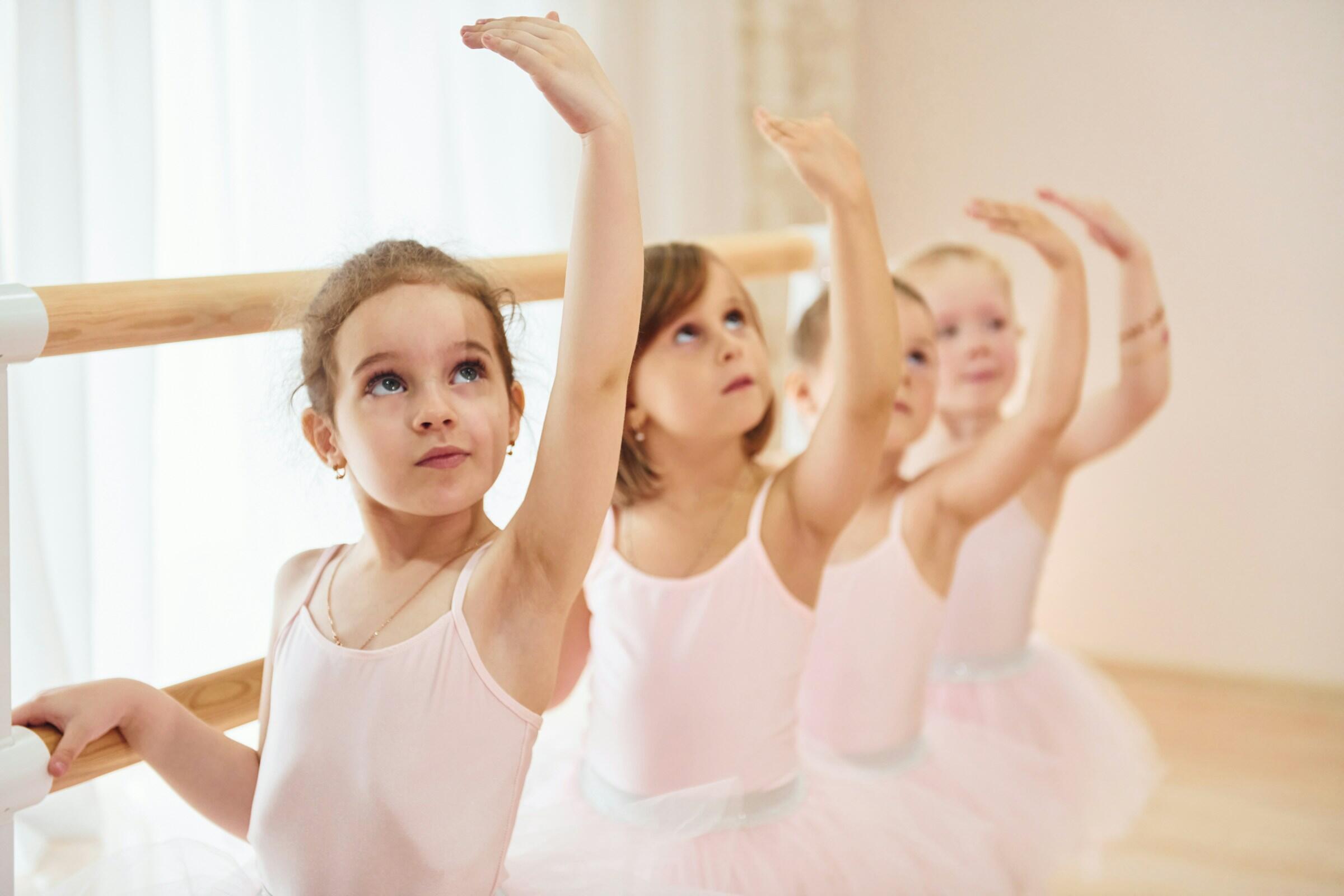Cuatro niñas pequeñas, futuras bailarinas, alineadas en una barra de ballet de madera en un estudio luminoso. Todas visten leotardos rosas y tutús blancos, con el brazo derecho levantado y la mirada hacia arriba en una pose de ballet.