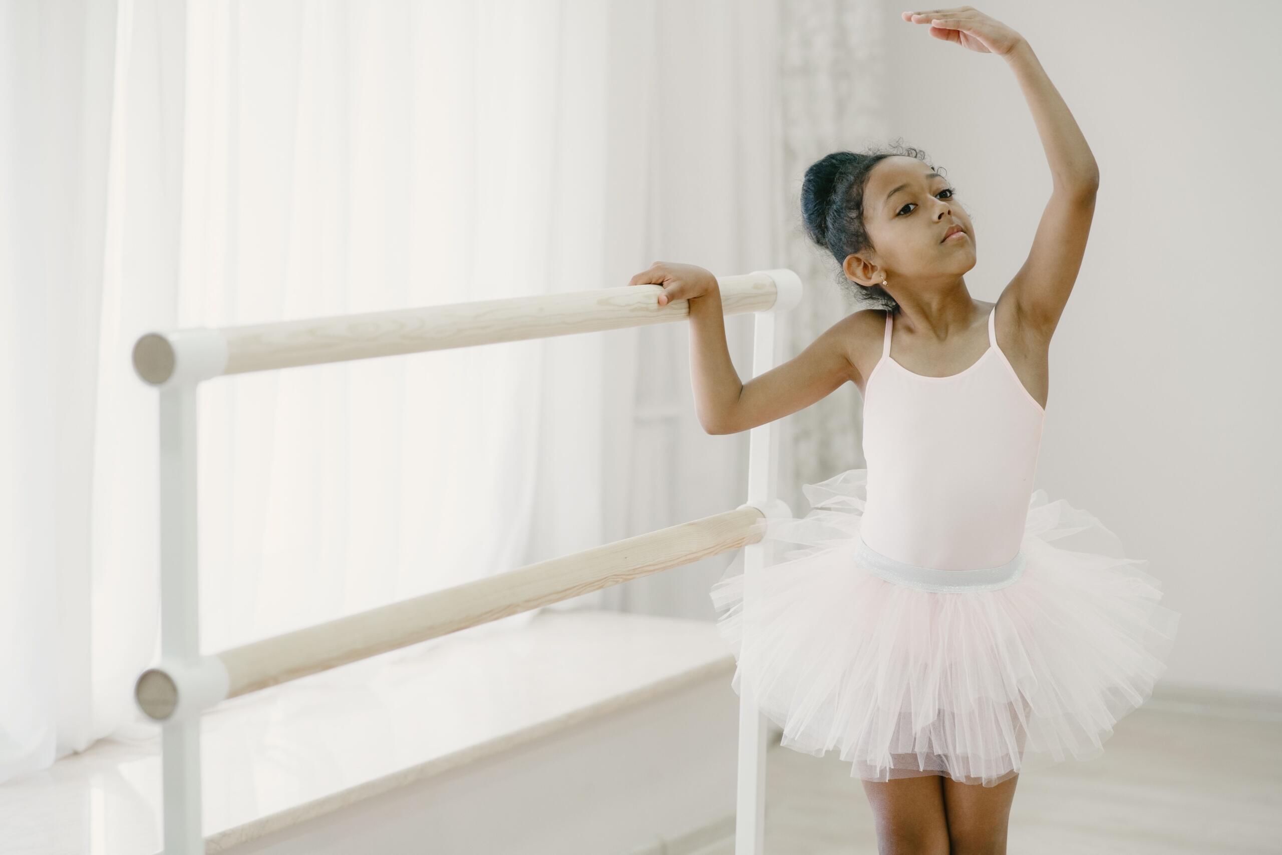 Una niña enfocada de ascendencia africana, con el pelo recogido en un moño, vistiendo un leotardo de ballet rosa y un tutú, se agarra a una barra de ballet de madera con una mano mientras levanta el otro brazo en una pose elegante y redondeada en una sala de estilo estudio bien iluminada.