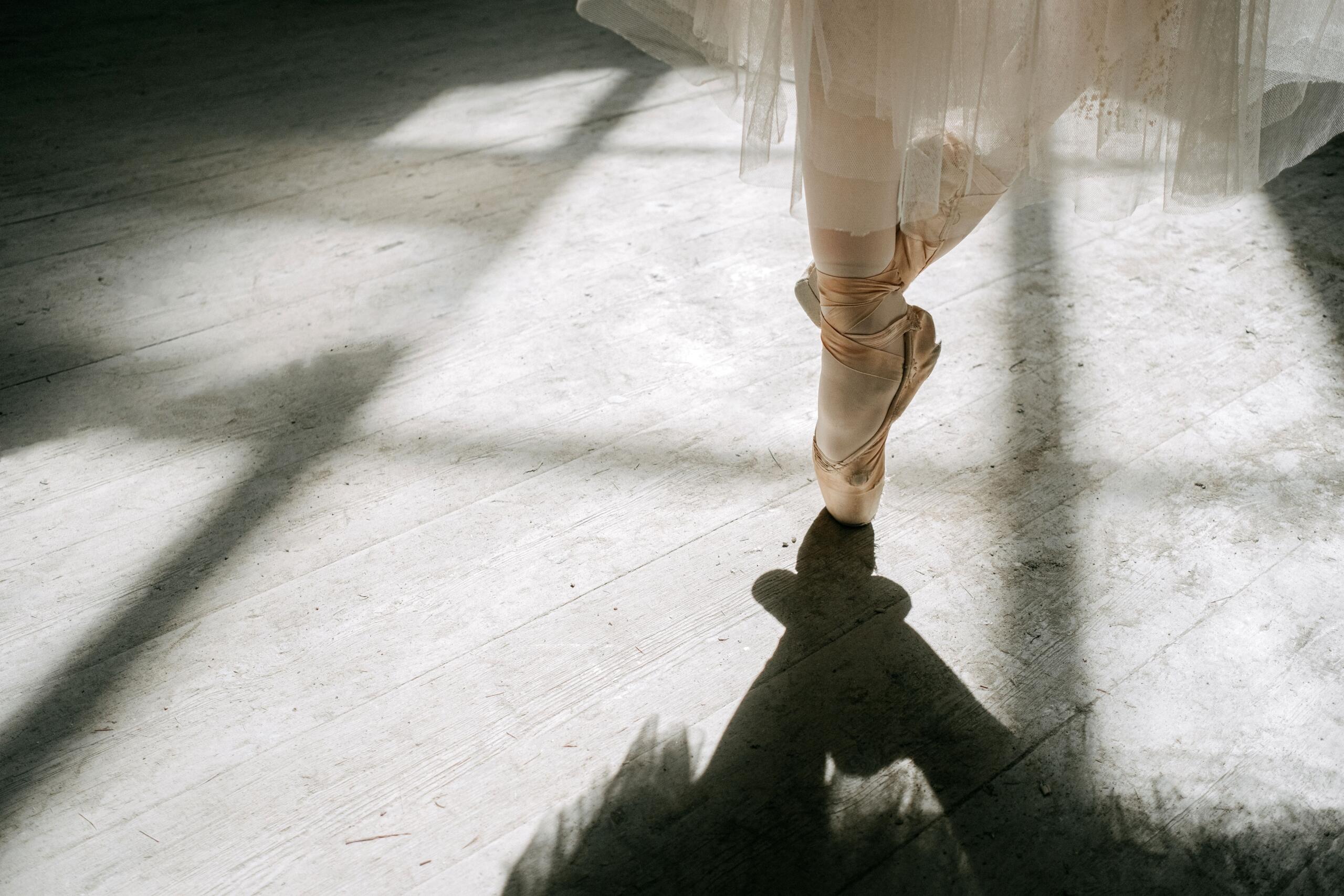 Plano detalle de los pies de una bailarina con tutú claro y zapatillas de punta de raso, manteniéndose en equilibrio de puntas (en pointe) sobre un suelo de madera donde la luz natural del sol proyecta sombras marcadas.