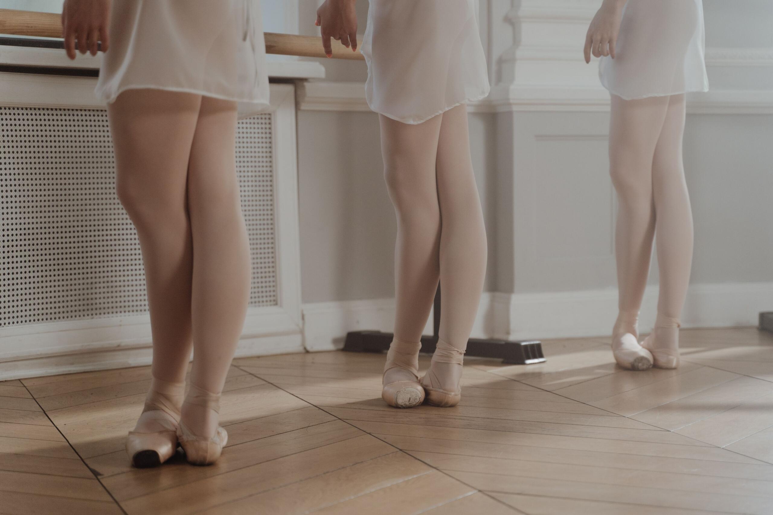 Plano enfocado en las piernas y pies de tres bailarinas de ballet con vestimenta blanca y zapatillas de punta, ejecutando la quinta posición de pies junto a la barra de ensayo sobre un suelo de parqué.