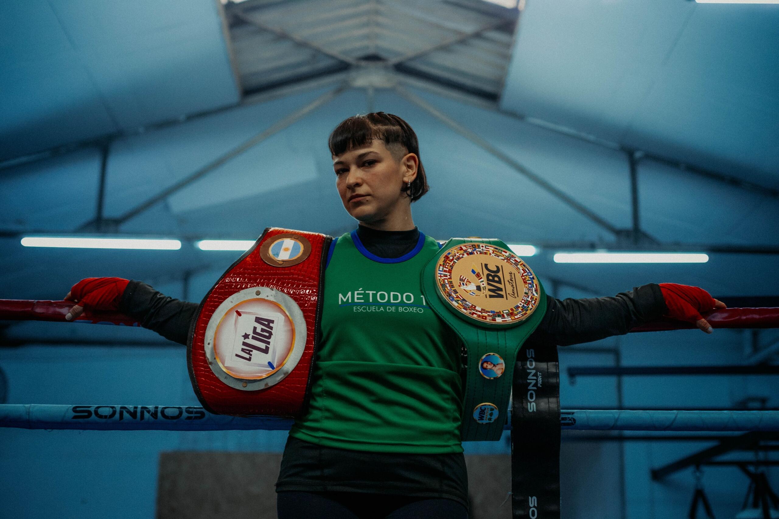 Boxeador de pie, con aire de confianza, en un ring, con dos cinturones de campeón colgados sobre una camiseta verde y el techo de un gimnasio de boxeo a sus espaldas.