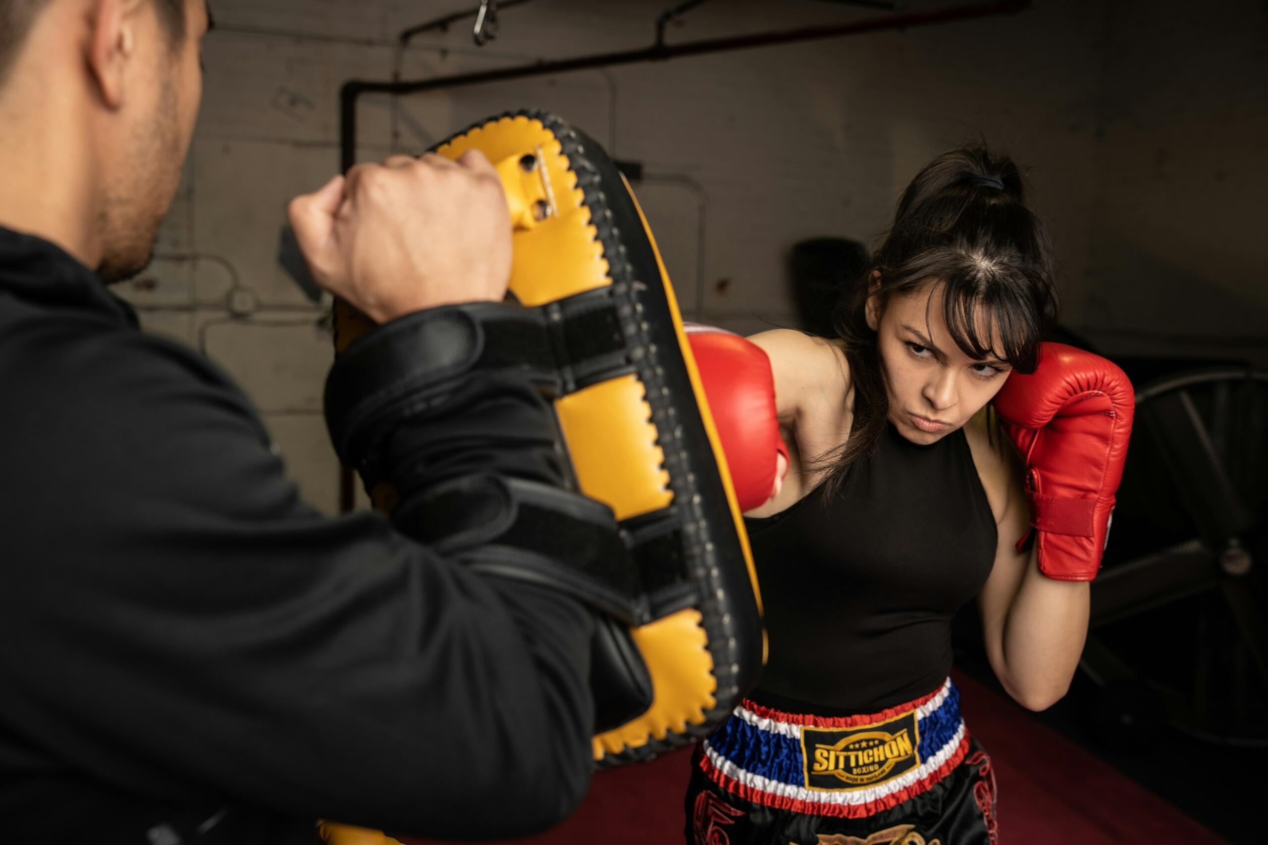 Una mujer con guantes de boxeo rojos que golpea un saco de entrenamiento amarillo que sostiene un entrenador en un gimnasio.