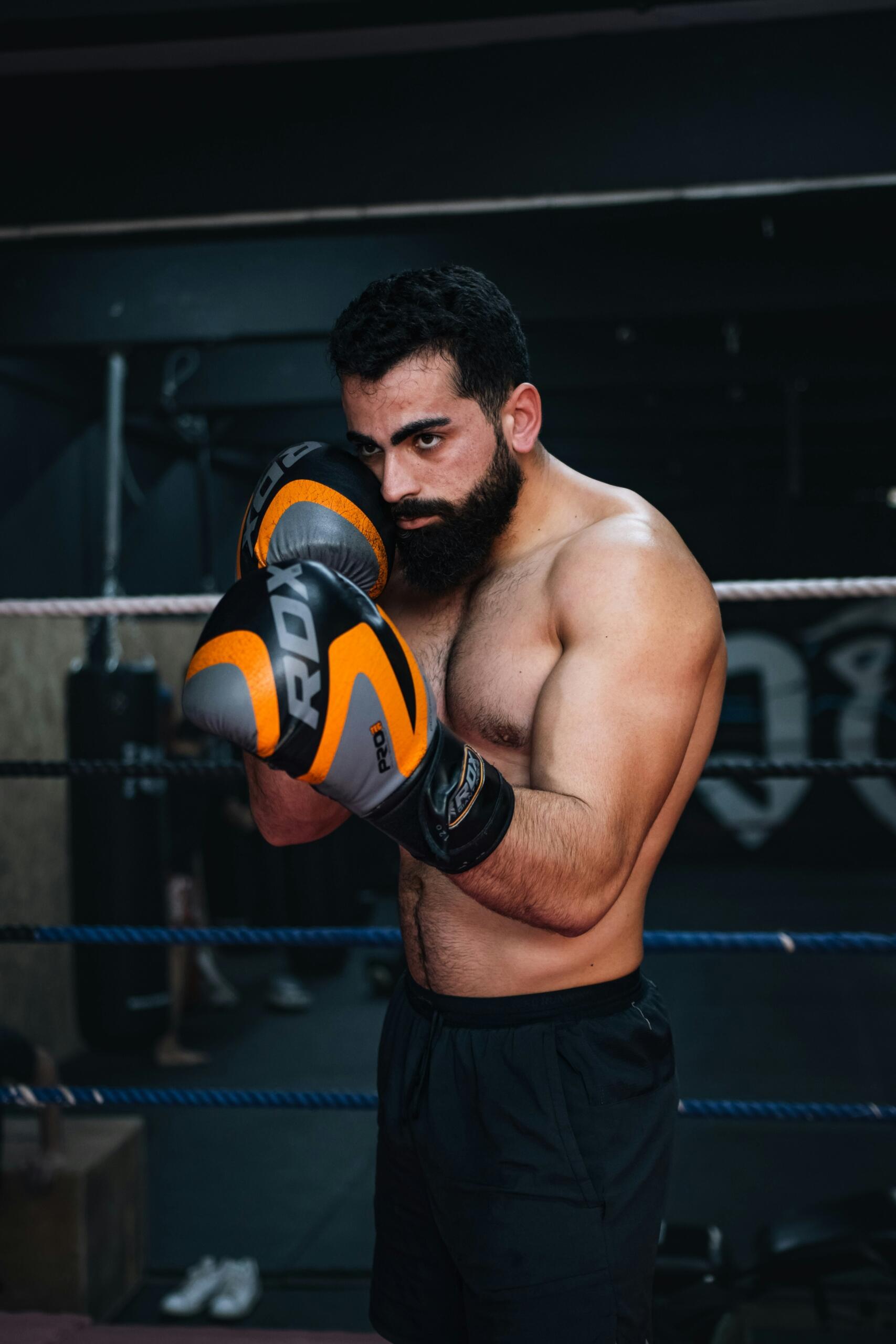 Hombre musculoso y barbudo, con pantalones cortos negros y guantes de boxeo naranjas, posando en posición defensiva dentro de un ring de boxeo con poca luz.