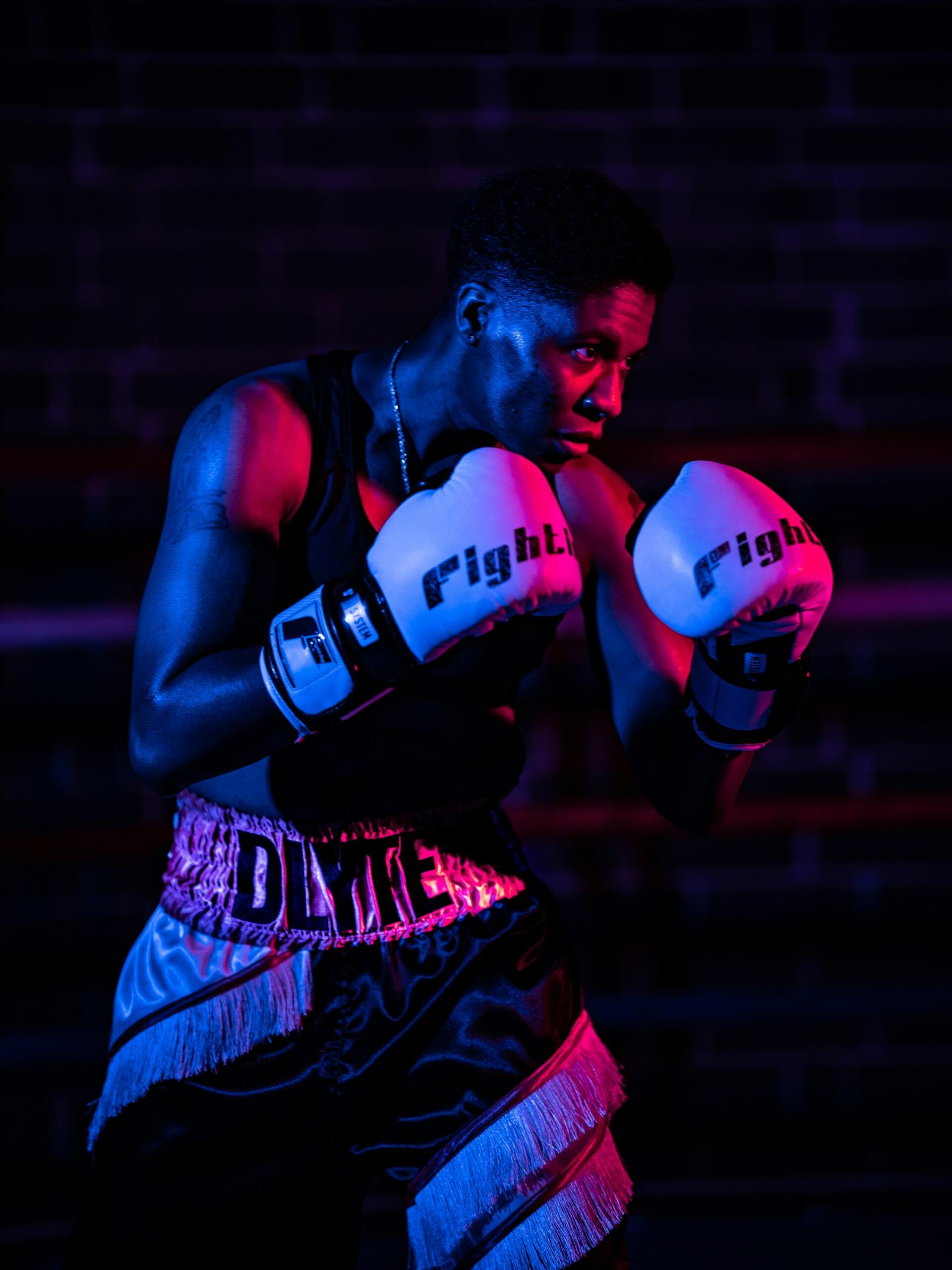 Boxeador en postura de combate, con guantes blancos y pantalones cortos negros, con luces de color azul neón y rosa sobre un fondo de ladrillo oscuro.