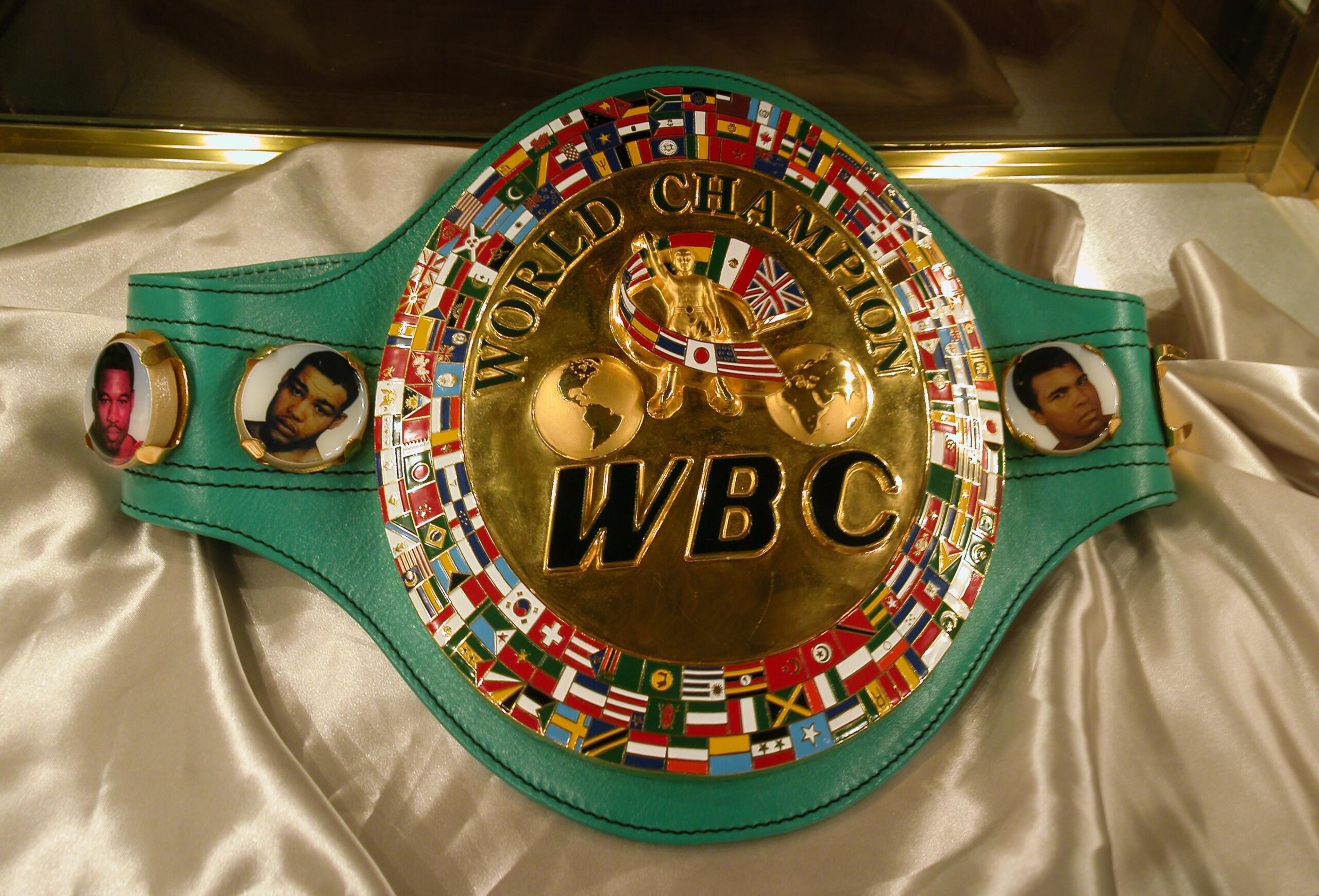 Cinturón de campeón mundial de boxeo de la WBC con placa dorada, un globo terráqueo y banderas sobre una correa verde, expuesto sobre una tela blanca.