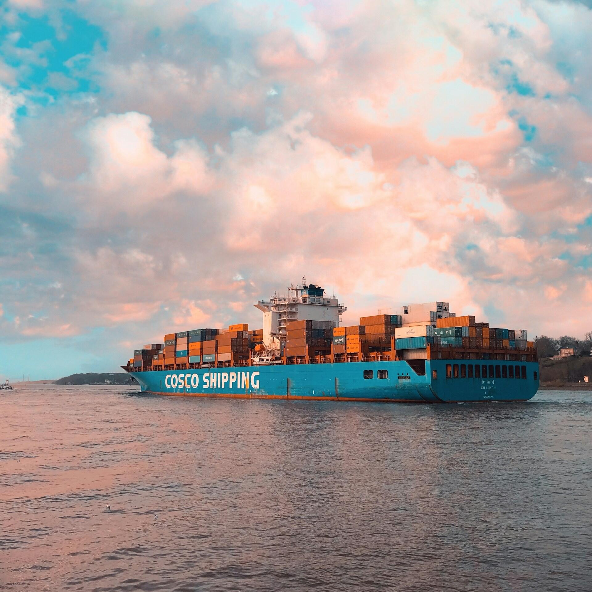Un buque portacontenedores de COSCO SHIPPING, de color turquesa y cargado de contenedores de colores, navega por aguas tranquilas bajo un espectacular cielo rosa y azul.