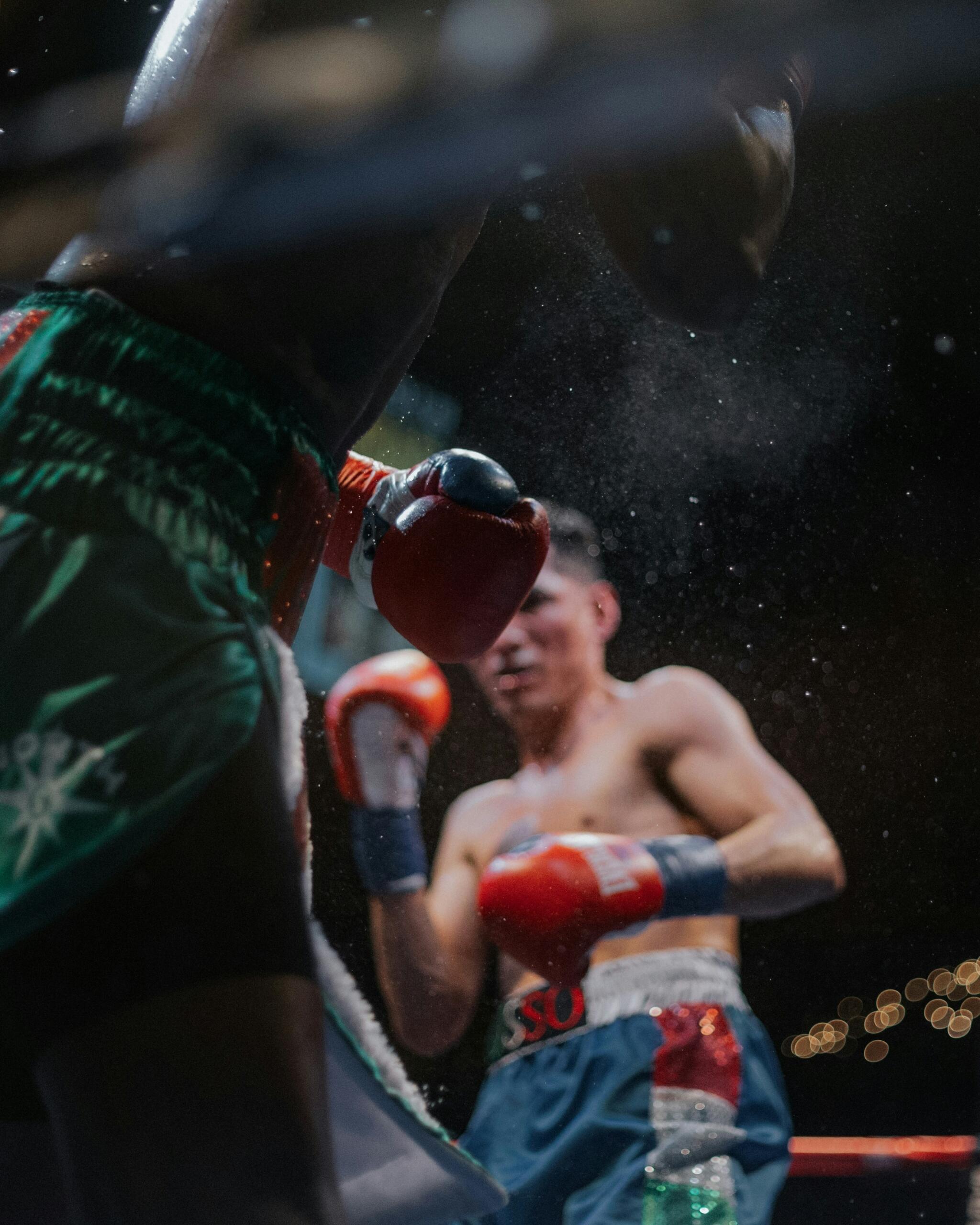 Dos boxeadores en un ring, uno de ellos parcialmente visible, intercambiando golpes con los guantes en movimiento y partículas de sudor o talco en el aire.