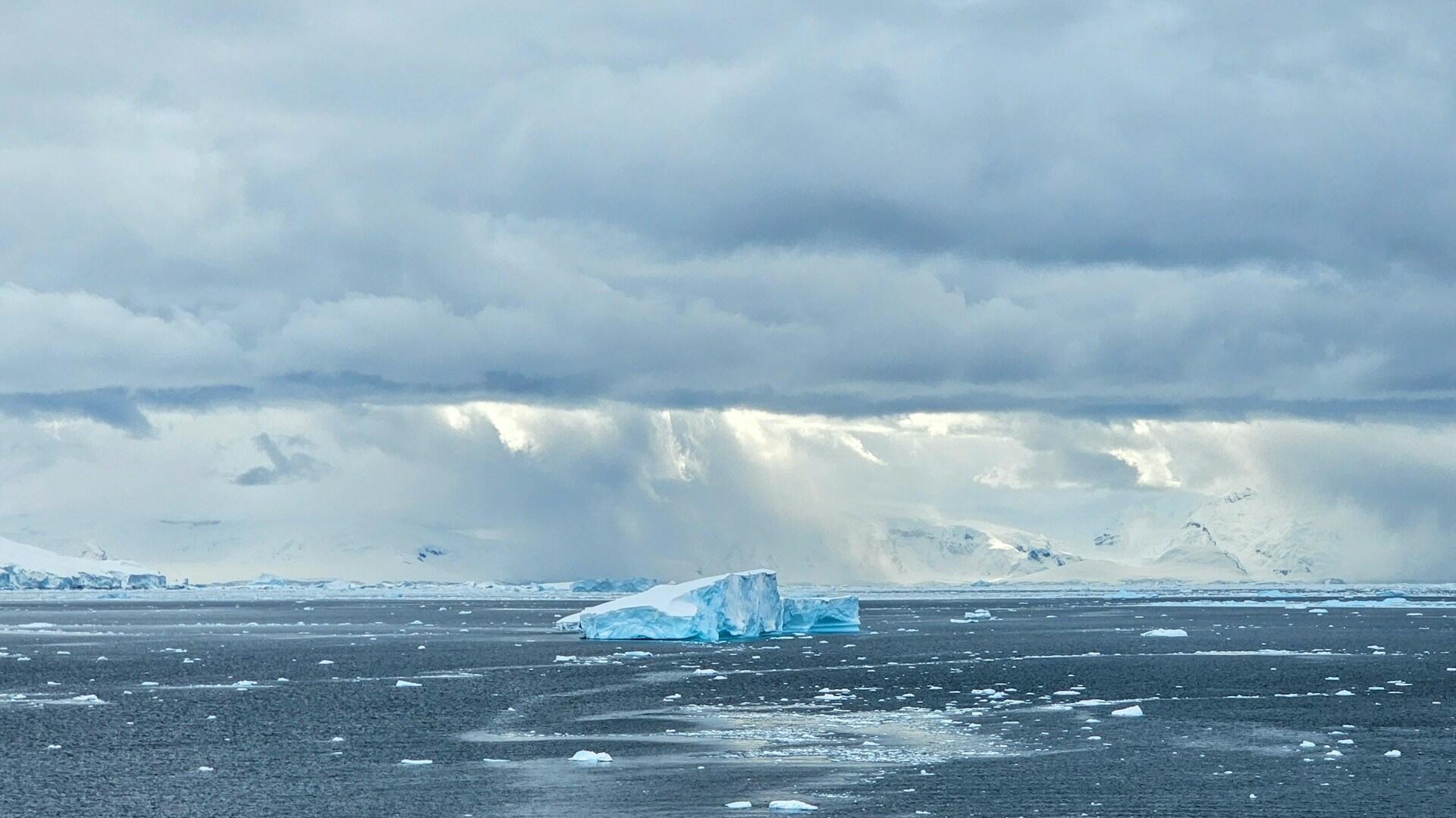 Un pequeño iceberg azul flota en un mar oscuro y helado bajo unas nubes de tormenta, con montañas nevadas en la lejanía y la pálida luz del sol en el horizonte.