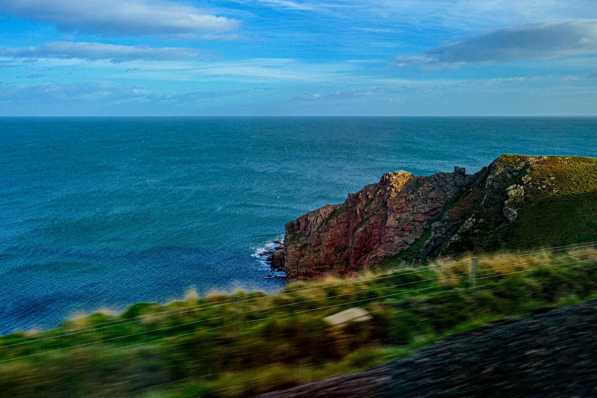 El paisaje costero, con un efecto de desenfoque por movimiento, muestra escarpados acantilados verdes y rojos sobre un océano de un azul intenso, bajo un cielo luminoso y nublado, lo que evoca una sensación de libertad.