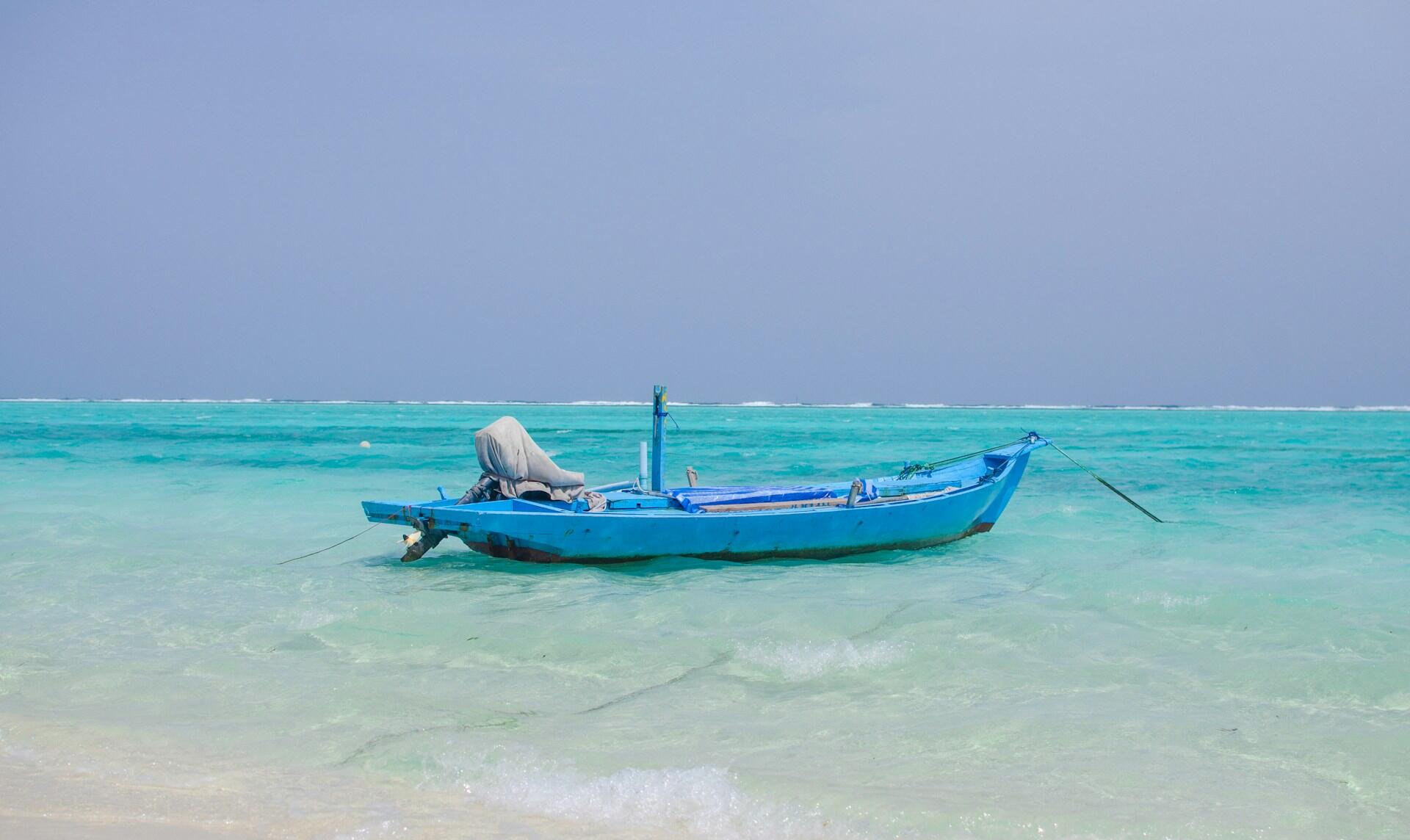 Un pequeño barco pesquero azul con redes a bordo flota en aguas poco profundas de color turquesa, anclado bajo un cielo azul y despejado; un sereno paisaje marino tropical.