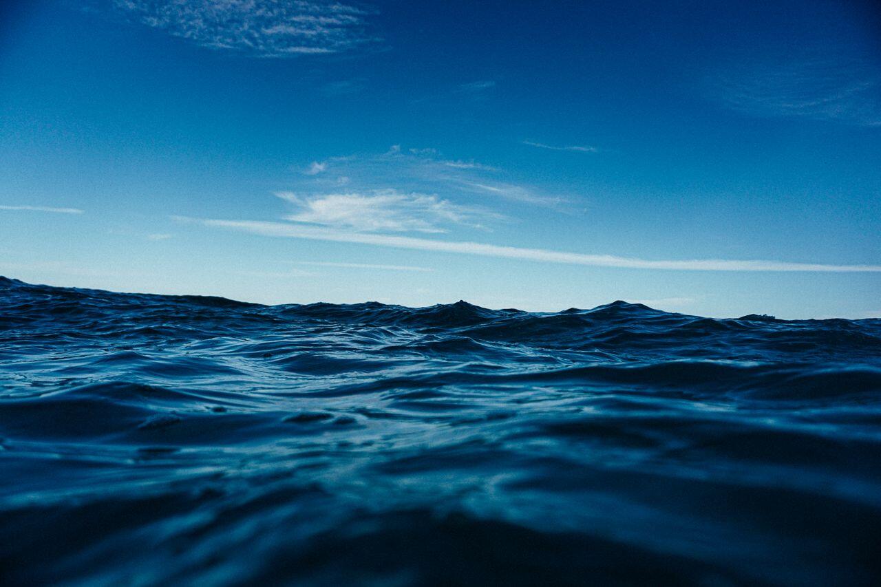Vista desde un ángulo bajo de las olas azul oscuro del océano bajo un cielo azul brillante, creando un paisaje marino tranquilo, amplio y ligeramente dramático.