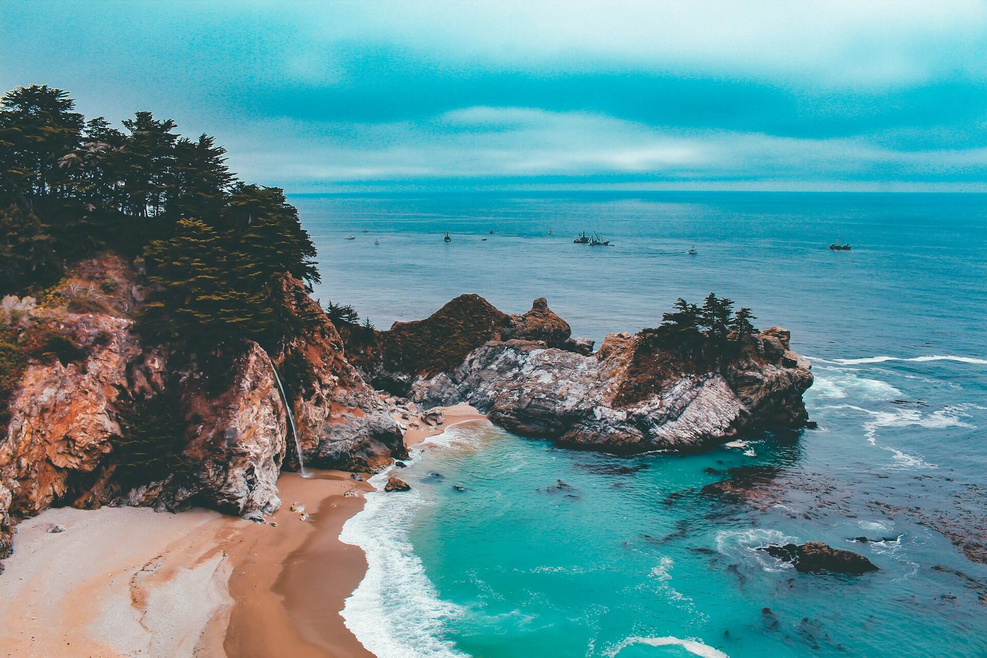 Las olas de color turquesa bañan una cala de arena bajo escarpados acantilados cubiertos de árboles e islotes rocosos; este paisaje costero, bajo un cielo nublado, transmite una sensación de serenidad y aislamiento.