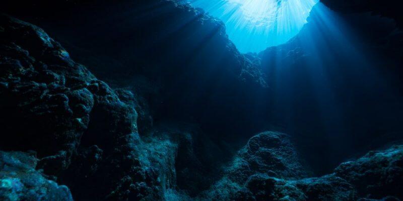 Una oscura caverna submarina con rocas escarpadas cubiertas de coral, iluminada por intensos rayos de luz azul que se filtran a través de la superficie, creando una atmósfera misteriosa.